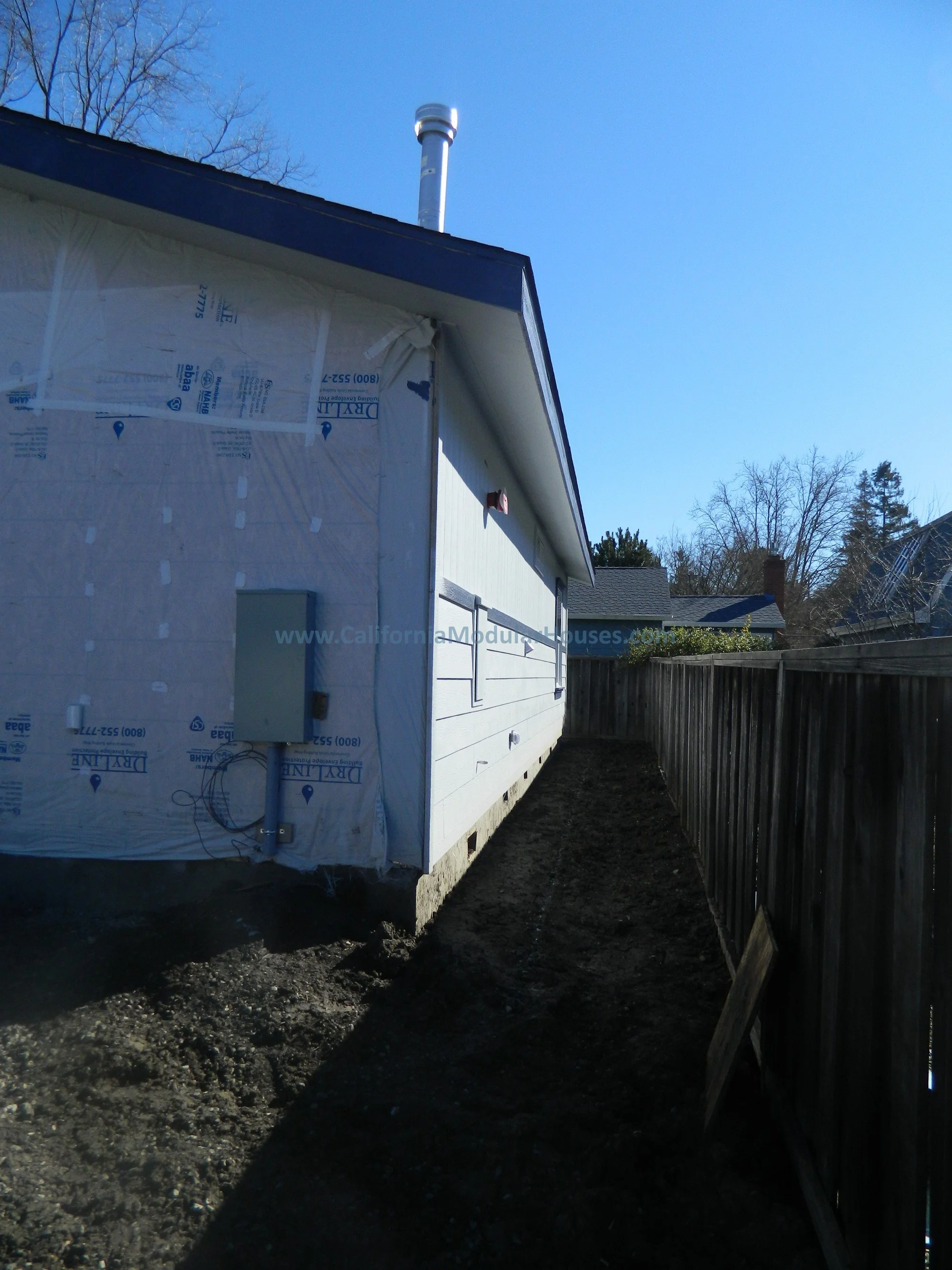 Side of a house under construction showing insulation on part of the exterior wall, a utility box, and a narrow dirt path alongside a wooden fence, with a chimney protruding from the roof against a clear blue sky.
