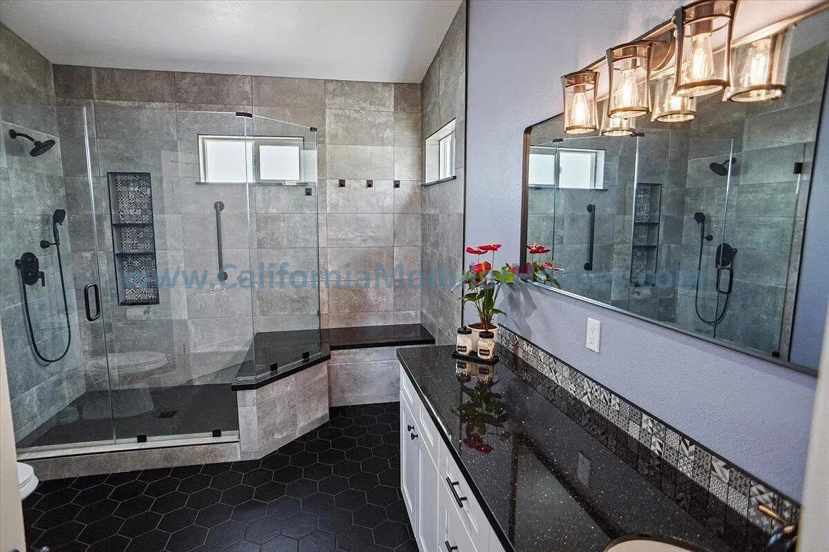 Bathroom with a large walk-in shower with glass doors, black fixtures, and small wall-mounted shelves, a black hexagon tile floor, a black granite countertop with a white cabinet below, a large mirror, and a light fixture with five glass lamps. There