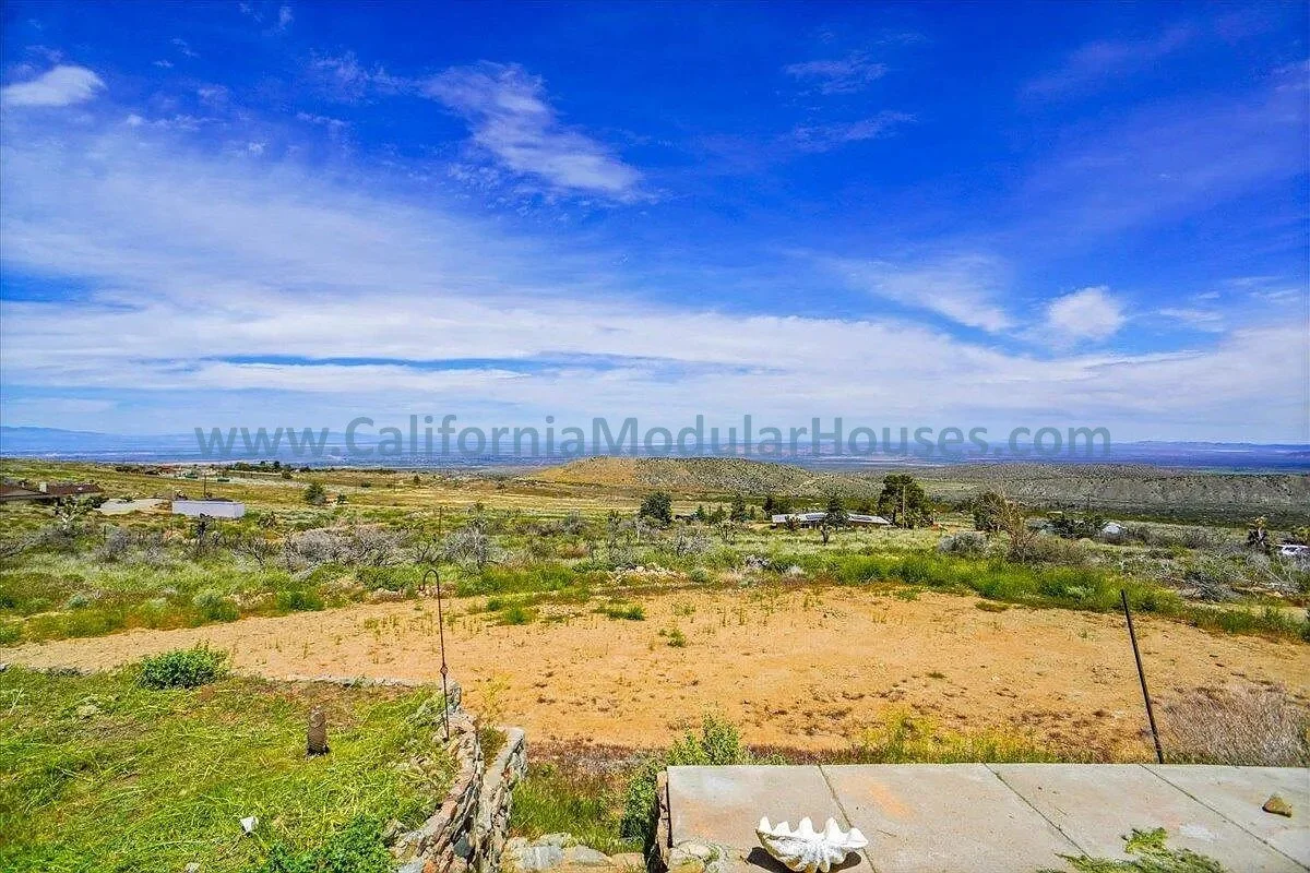 A wide landscape view of a desert area with sparse vegetation, some small structures, and an expansive blue sky with clouds.