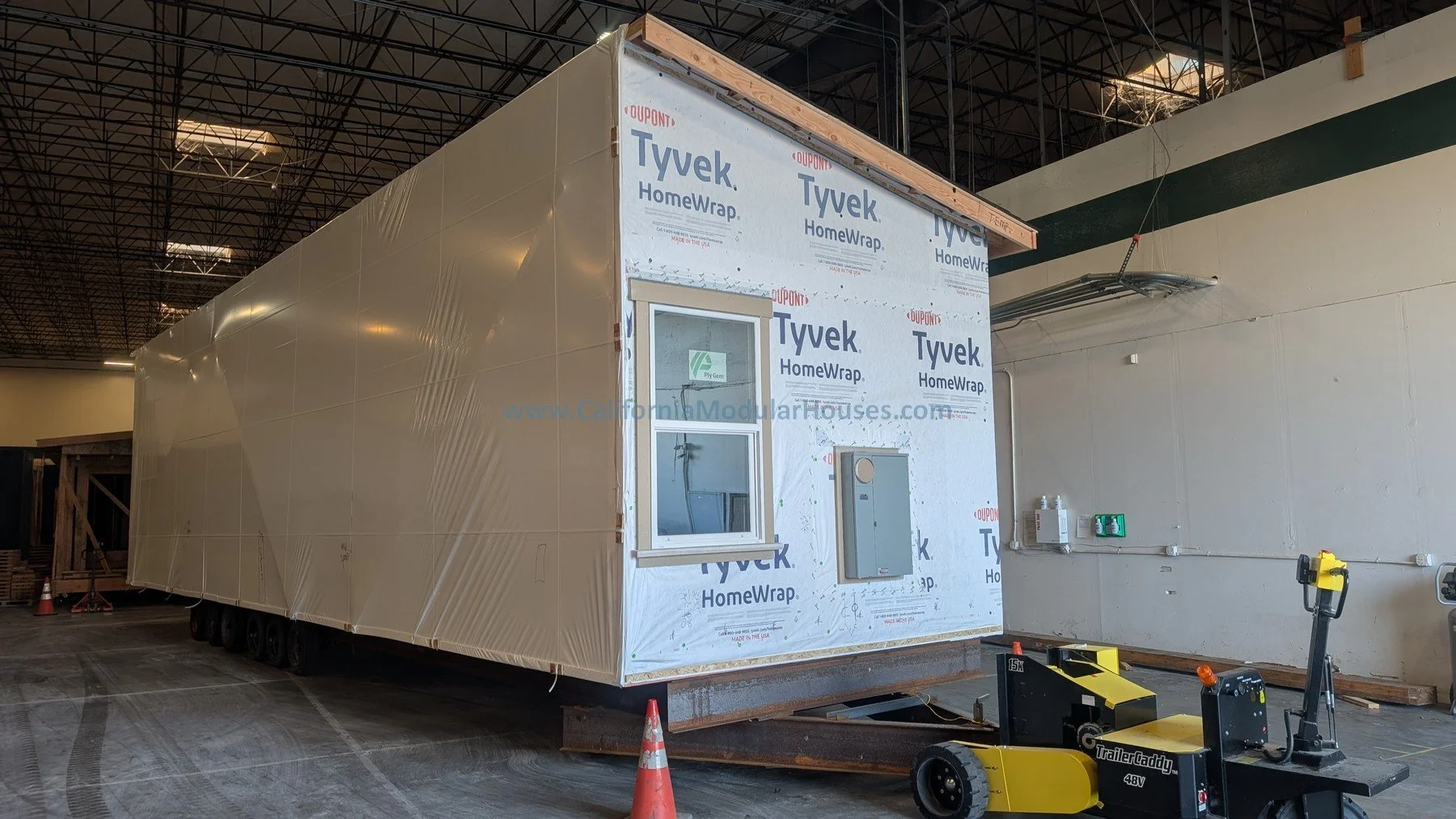 Construction site inside a warehouse showing a partially built modular house with Tyvek house wrap, a window, and electrical boxes.   Prefab modular home.  ADU. Accessory Dwelling Unit.  California.  