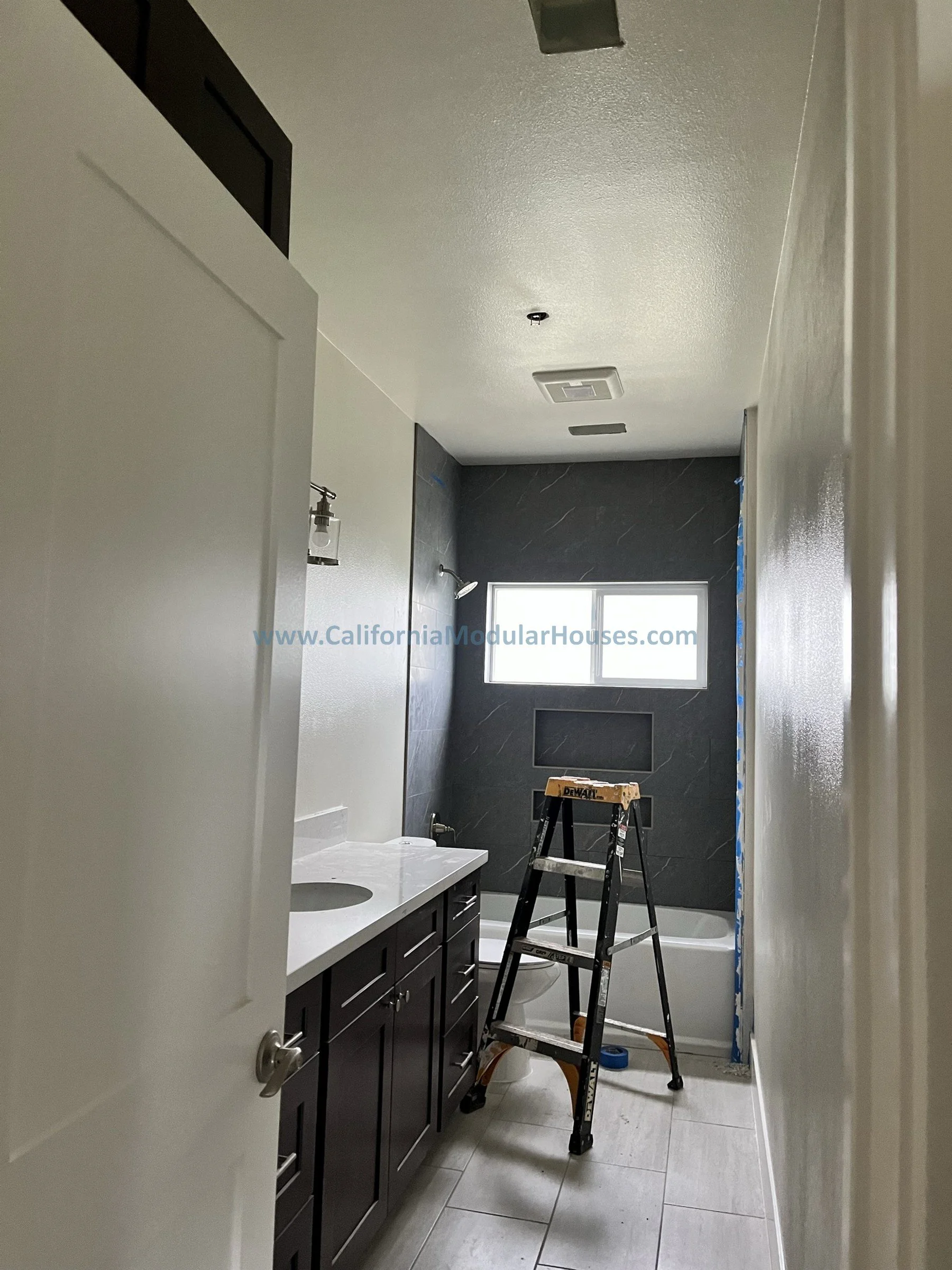 Bathroom under construction with dark wall tiles, a window, a partially installed bathtub, a dark vanity with a white countertop, and a stepladder.
