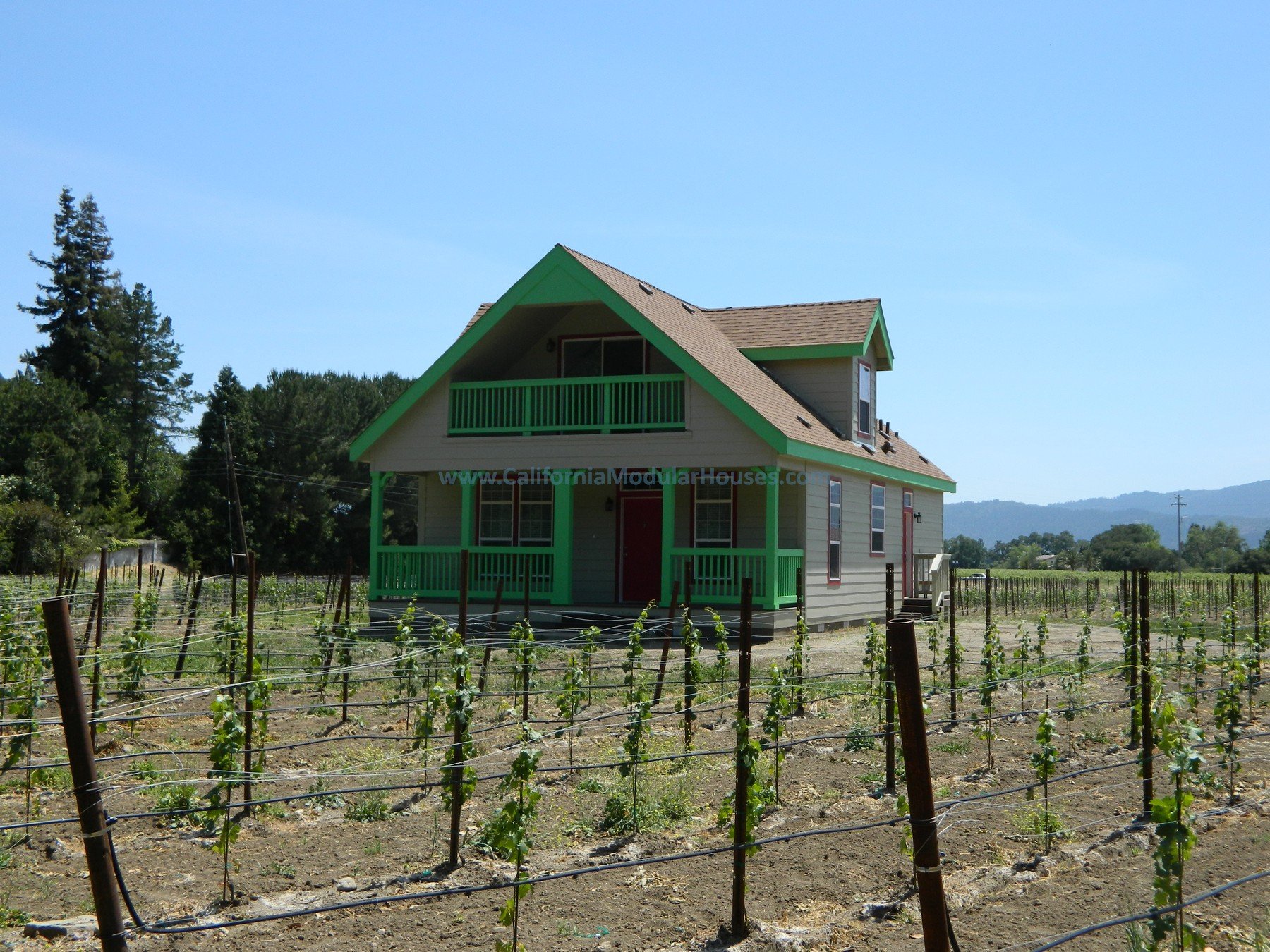 A two-story house with a green balcony and trim, beige siding, set in a rural area with vineyard rows in the foreground and mountains in the background.
