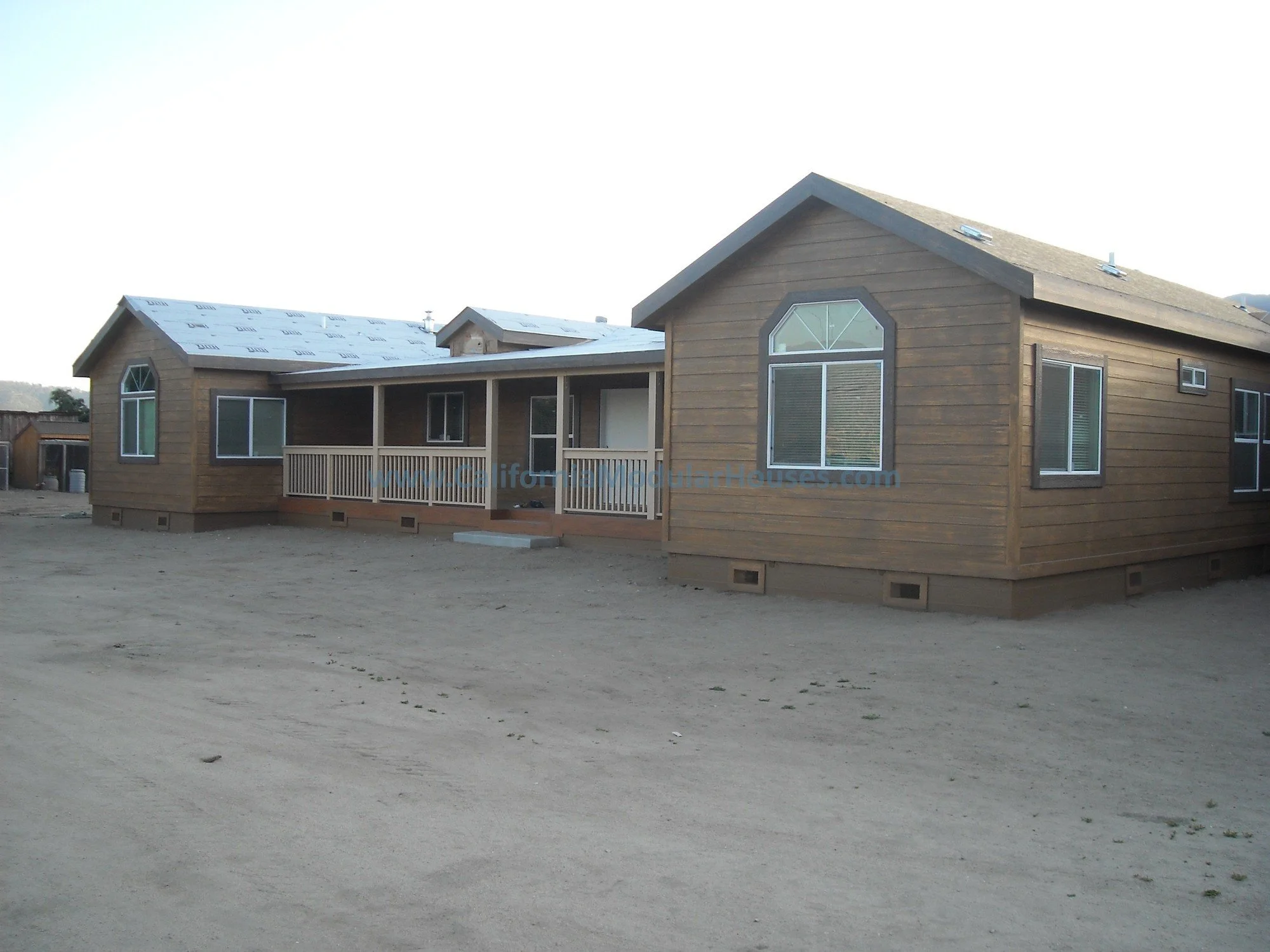 A new wooden house under construction with windows installed and a porch, in a rural area with dirt ground.