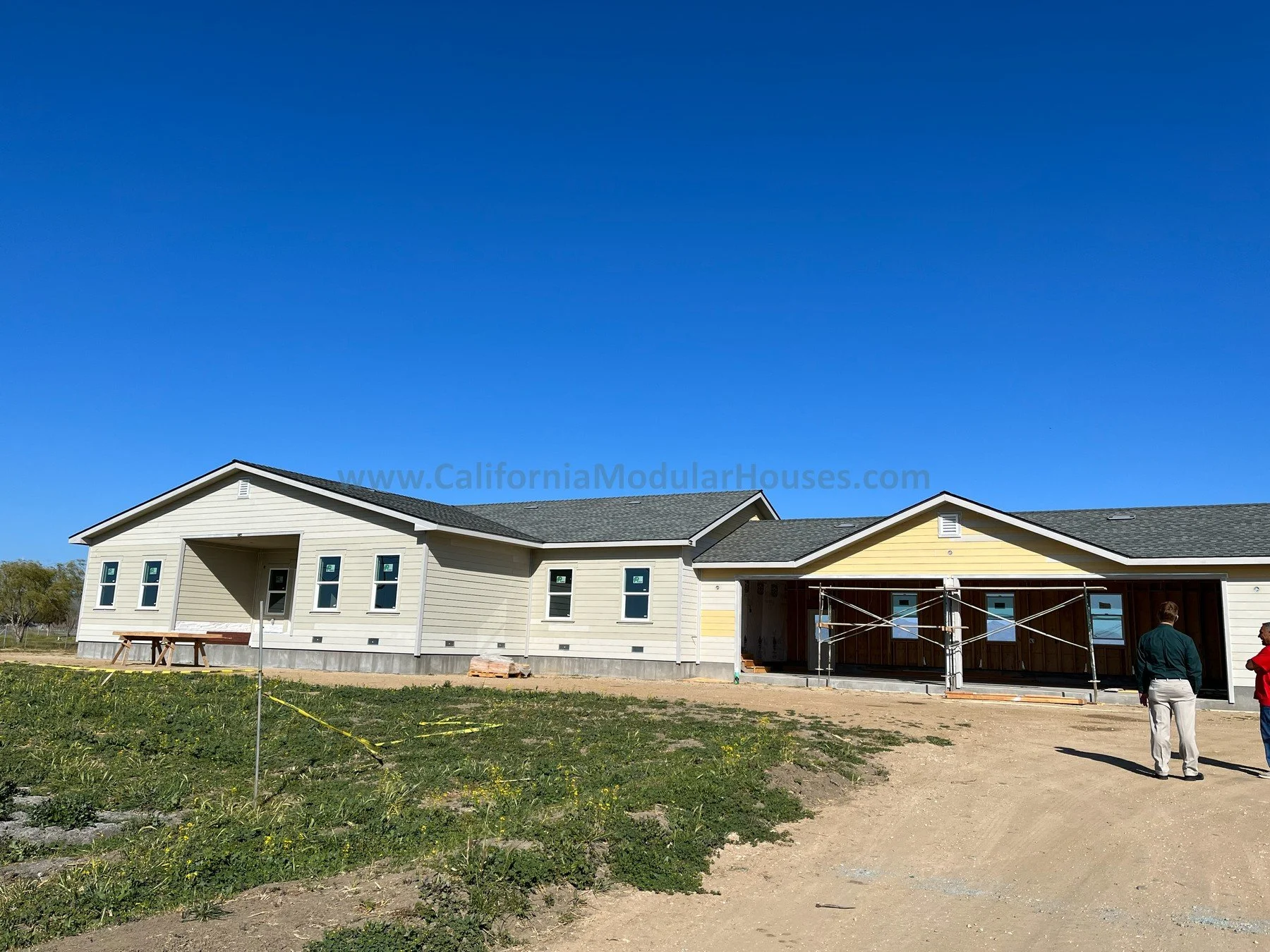 Construction site of a modern house with partly installed siding, two people standing on a dirt pathway, and a clear blue sky.