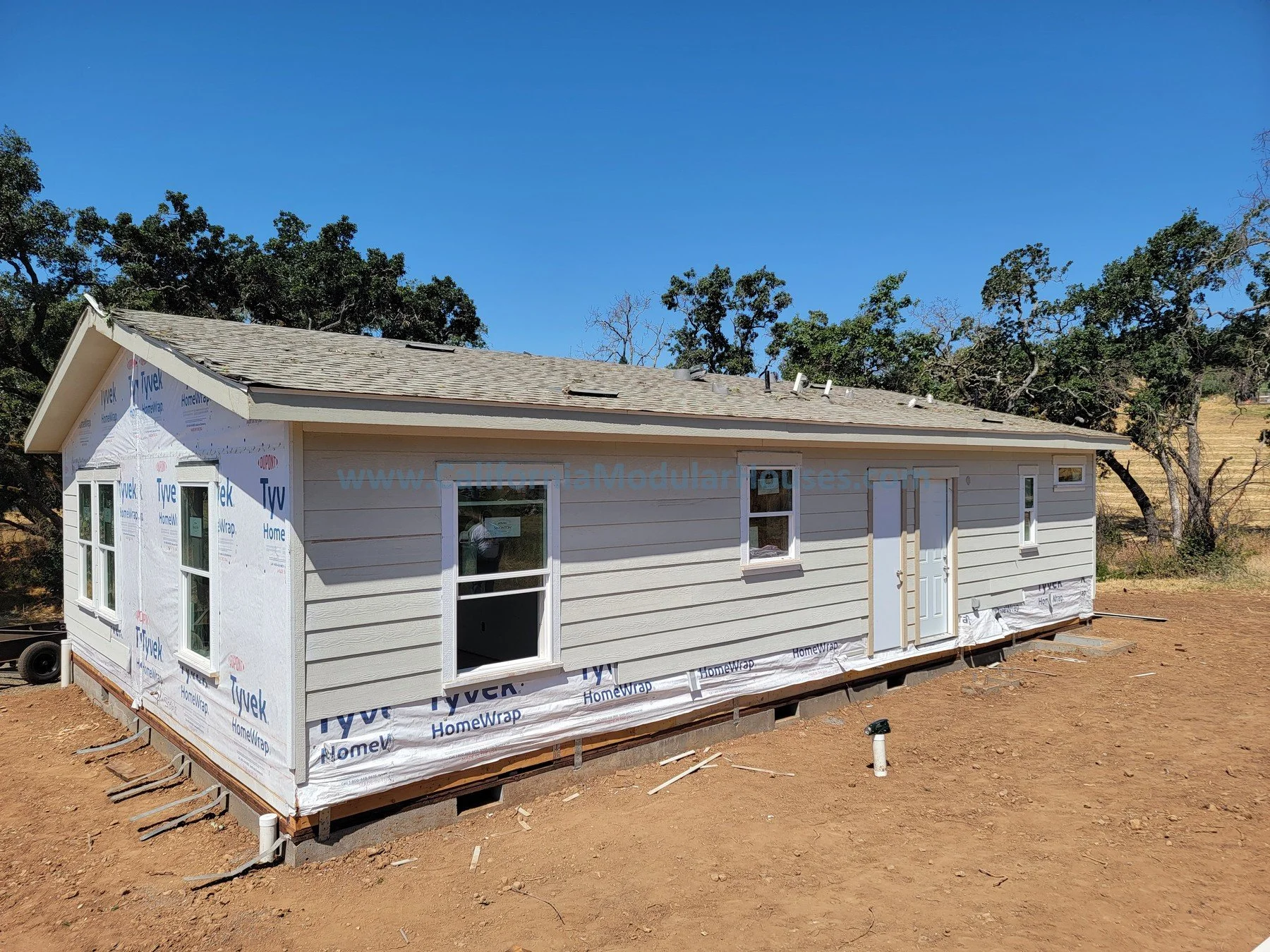 A house under construction with vinyl siding partially installed, numerous windows, and a white door, set on a dirt lot with trees in the background and a clear blue sky.