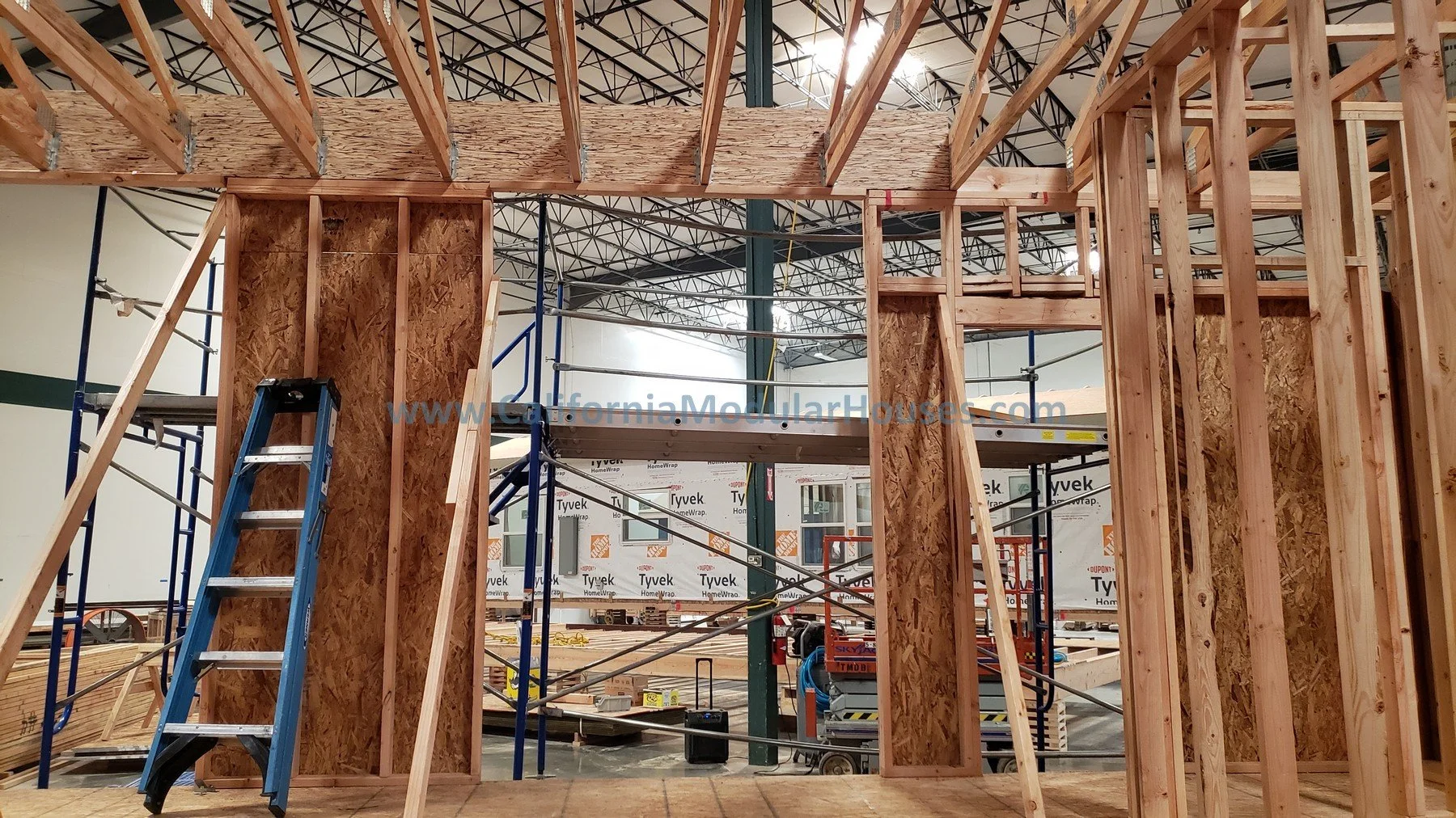 Interior of a building under construction showing wooden framing, a blue ladder, and building materials, with a look at the ceiling's metal support beams.