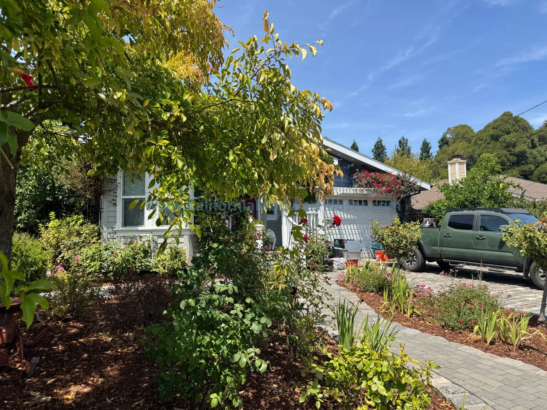 A residential house with a garden in front, flowers, and a parked gray truck on a driveway. Trees and shrubs surround the house, with a clear blue sky overhead.