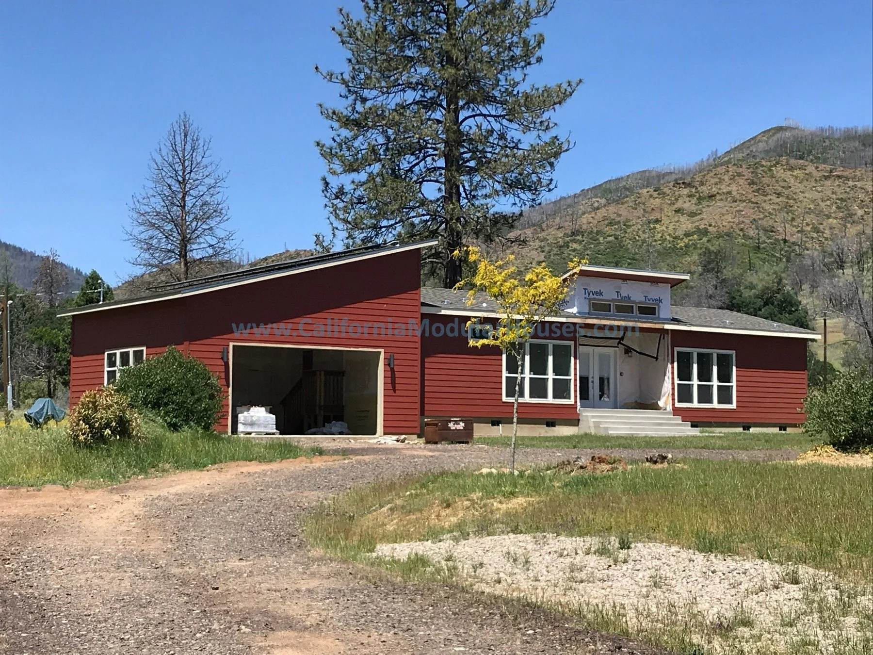A red modern house with a garage, white window frames, steps leading to front door, and a small tree in front.  Modular Homes in California, Pre-Fabricated Homes.
