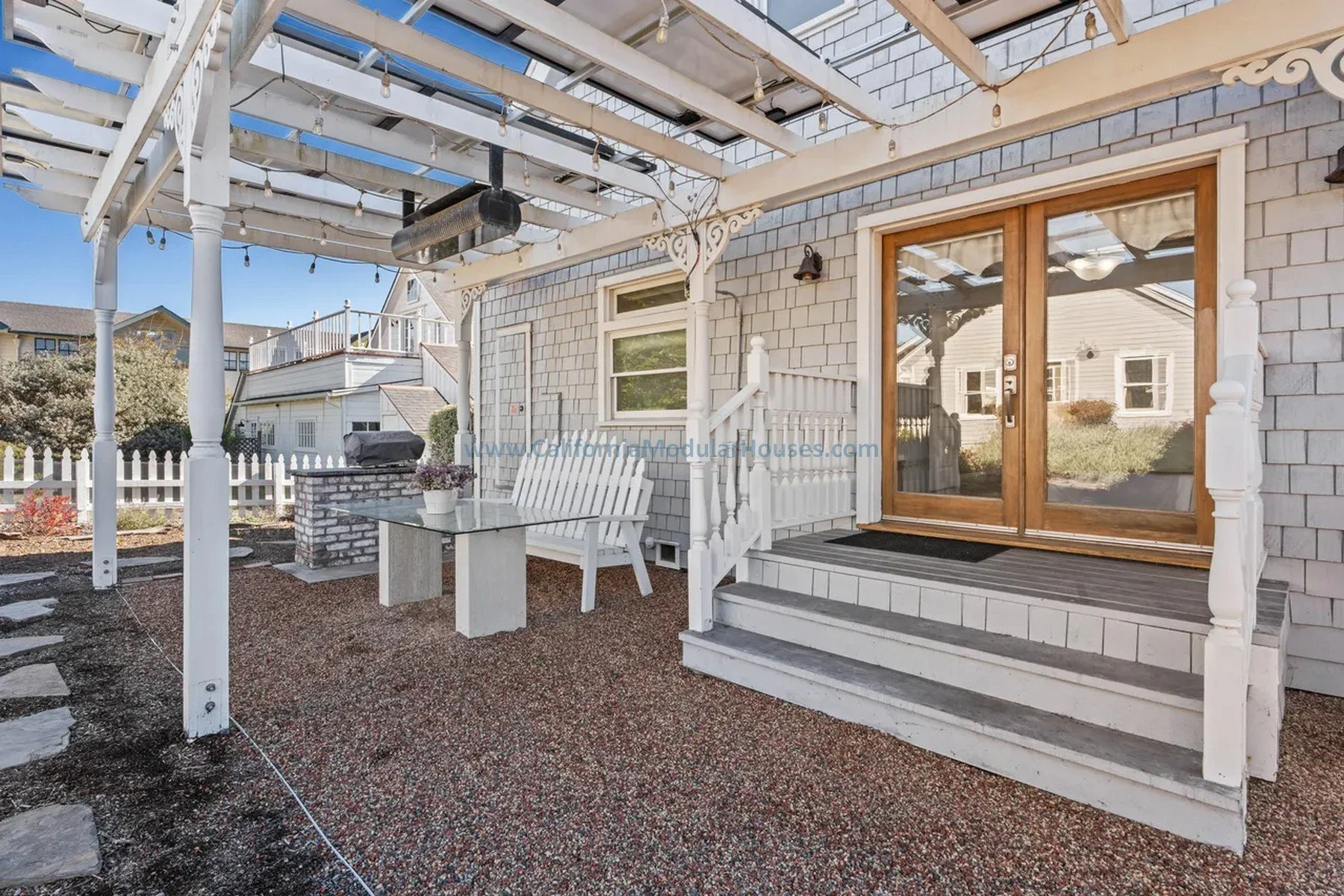 Cape Cod home with attic finished on-site.  California.  Modular prefab house.  Backyard patio with white lattice pergola, glass table, white wooden bench, potted plant, sliding glass door with wooden frame, stucco house exterior, steps.