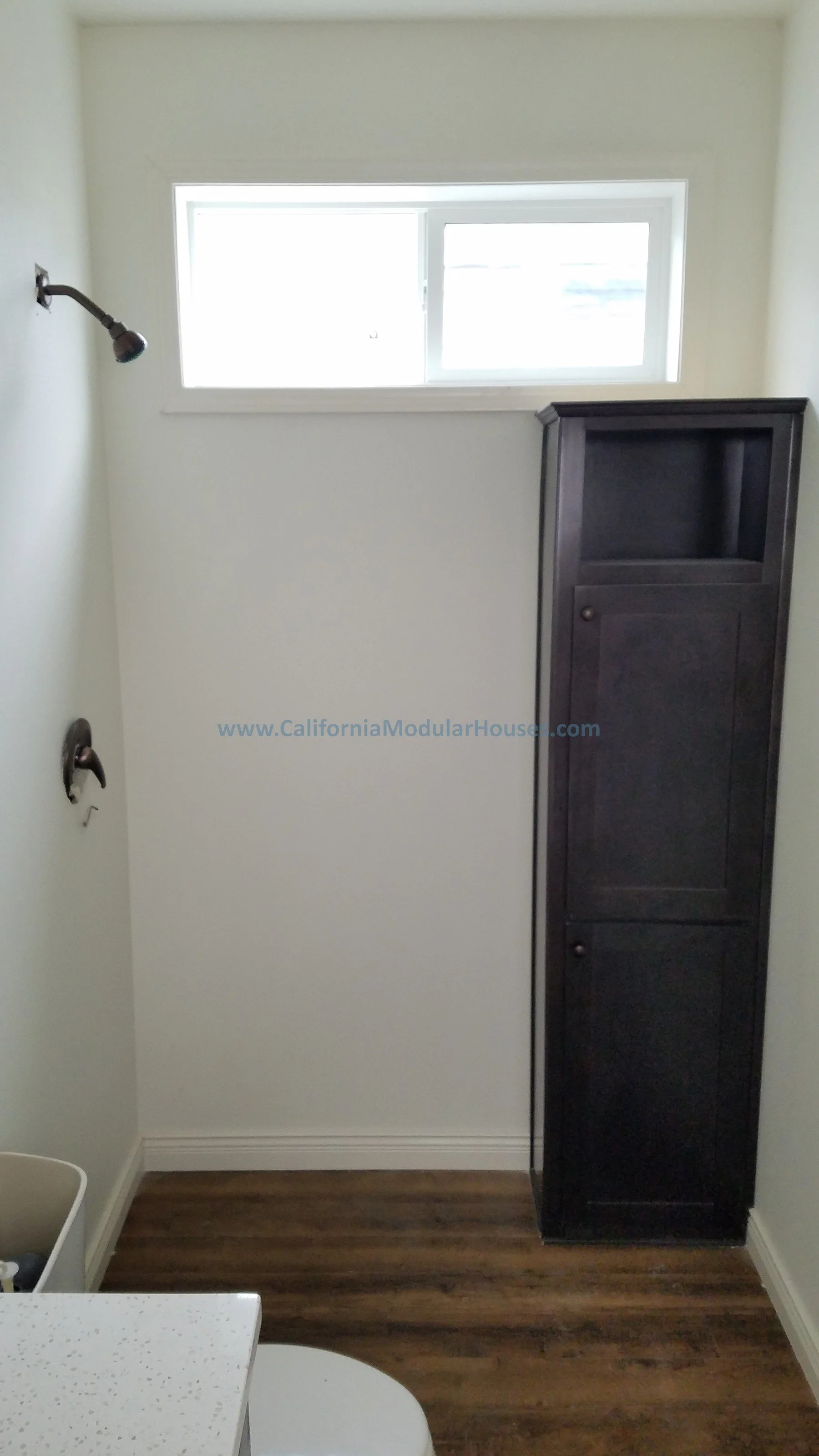 Empty bathroom with a showerhead, faucet, and a dark wooden cabinet next to a small window.  Pre-Fabricated Homes, Prefab Home California, Modular Homes in California,