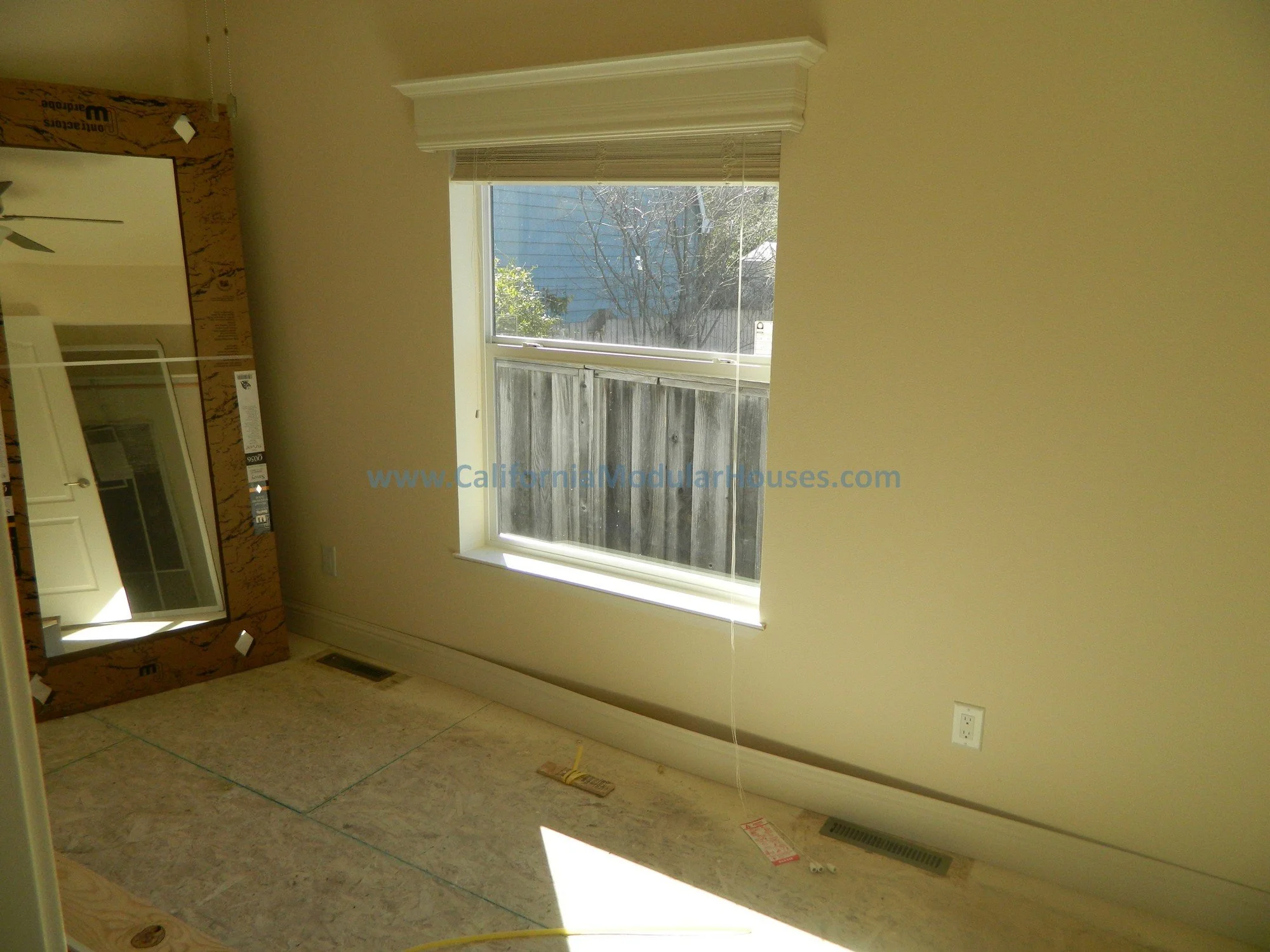 Empty room with beige walls, a window with blinds, a mirror leaning against the wall, and construction materials on the floor.