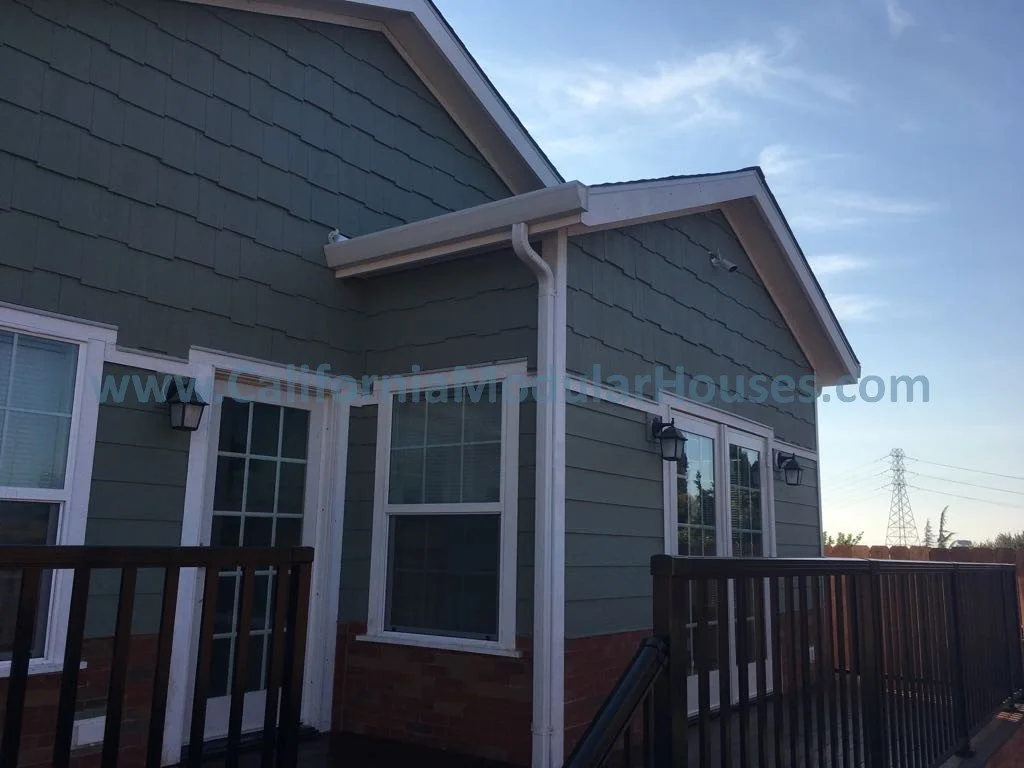 The back exterior of a house painted green with white trim, featuring multiple windows, a small porch with a black railing, outside wall-mounted lamps, and a white rain gutter system.