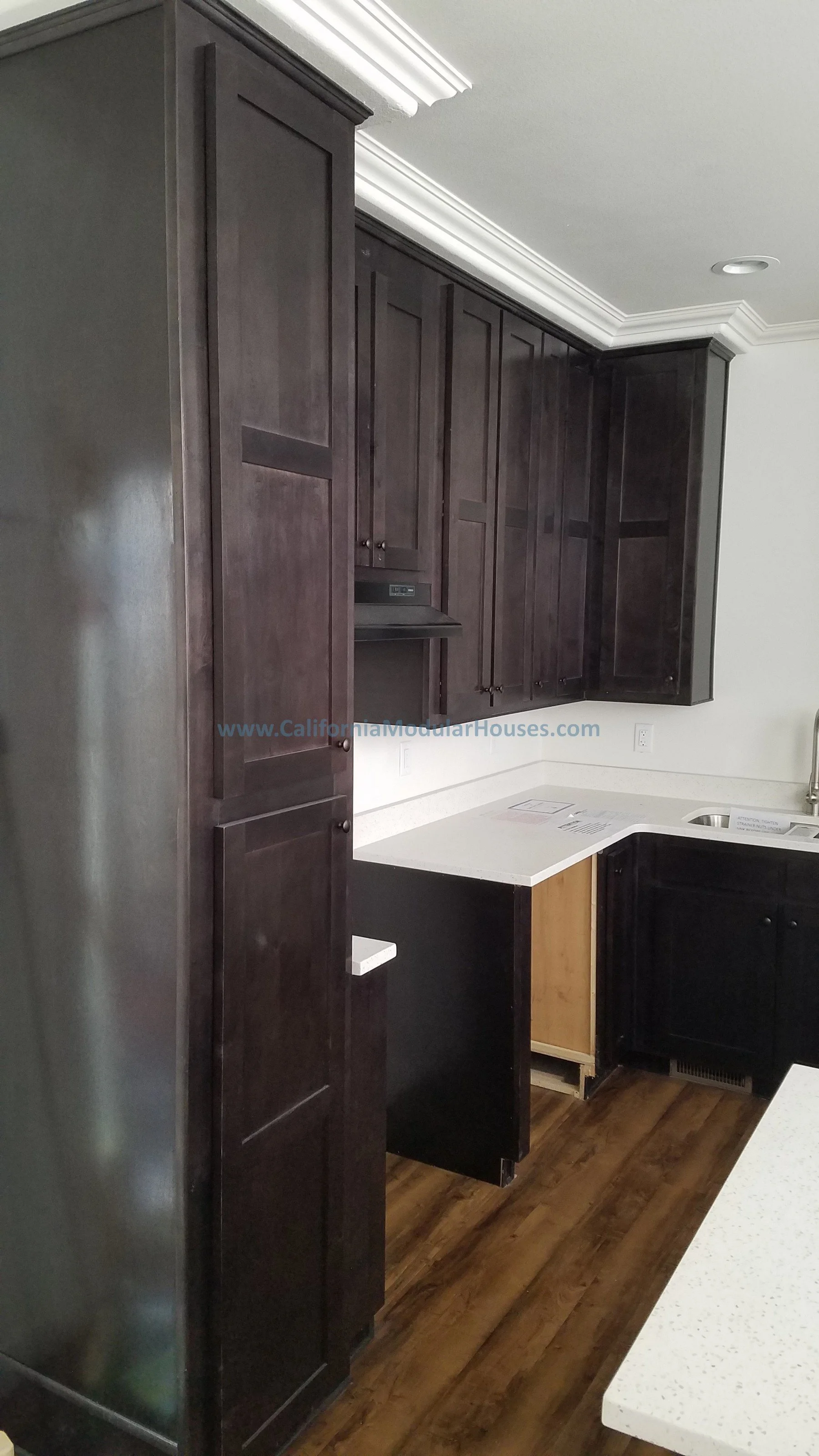 Kitchen with dark wood cabinets, white speckled countertops, a stainless steel sink, and wood flooring being completed in the factory.  Modular Home CA, California Modular Homes, Prefab Modular Homes California,