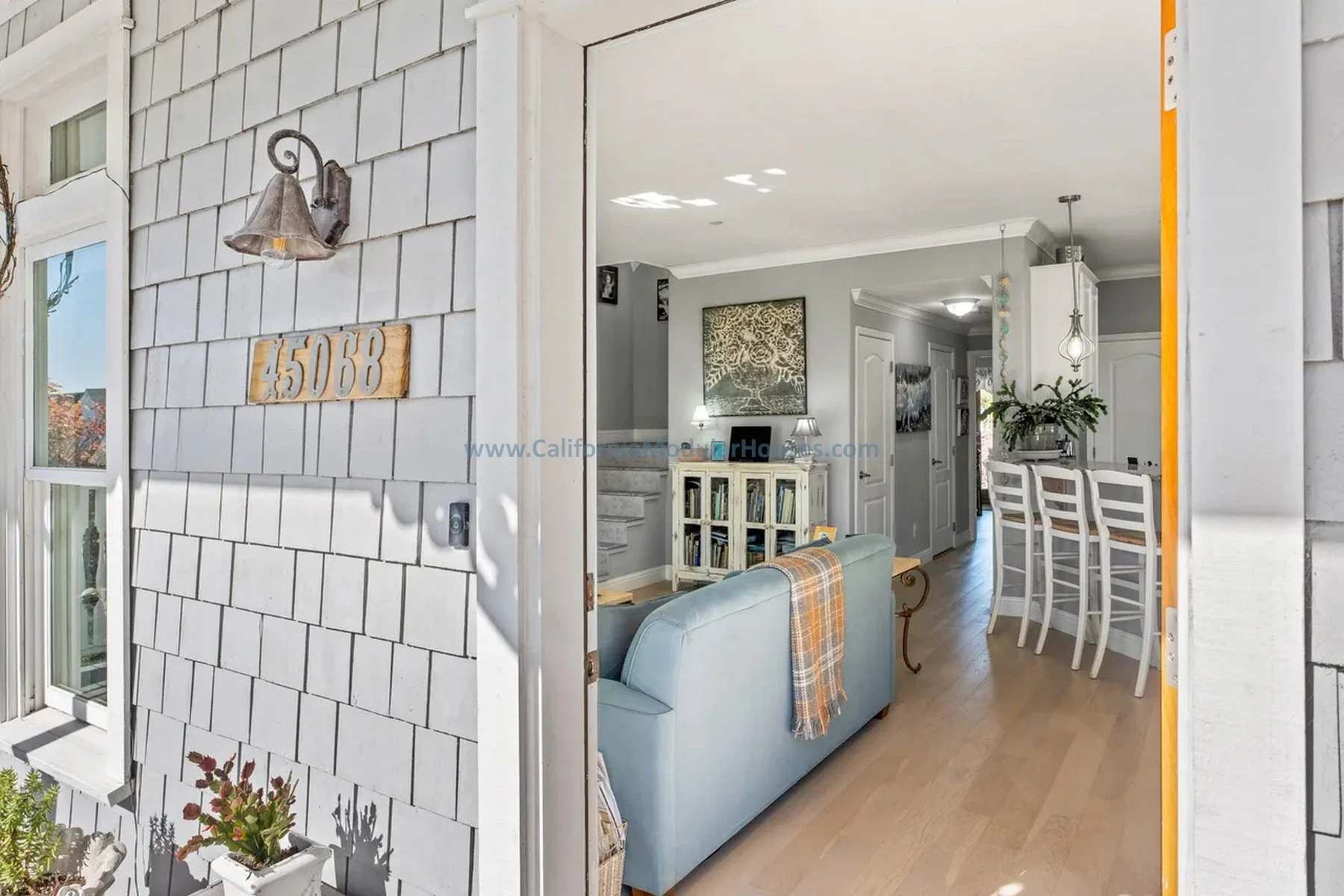 California. Pre-fab modulars. Custom design coastal commission cape cod modular home.  View from the front door into a living room and dining area of a house, painted shingle siding, and sunlight casting shadows.