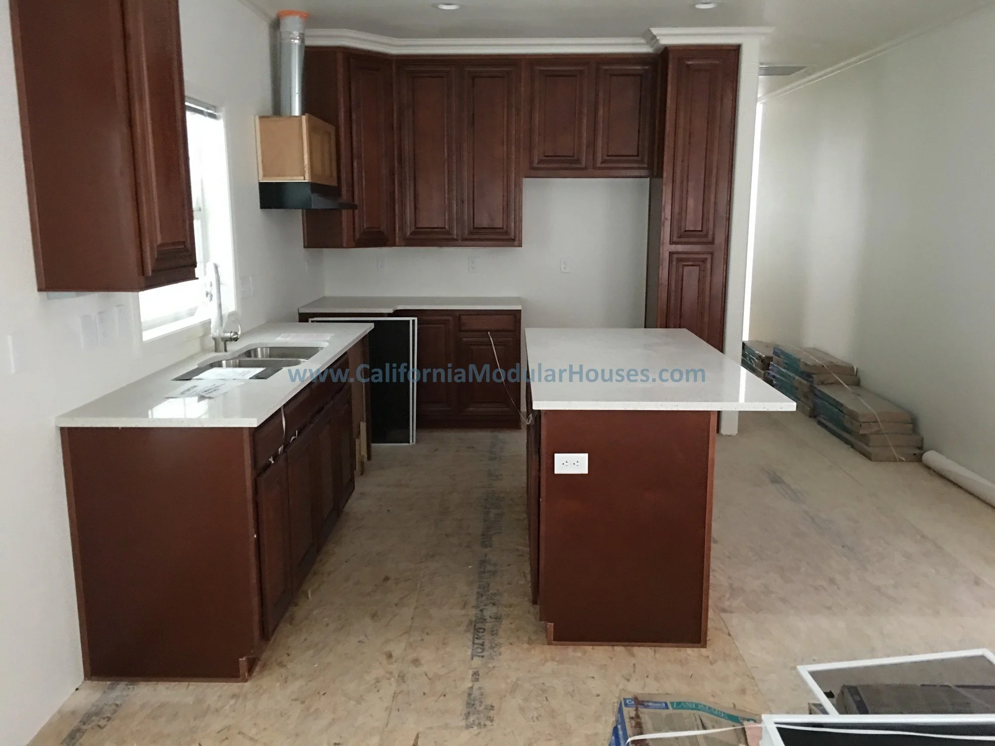 Kitchen with dark wooden cabinets, white countertops, a small sink, and an island counter. There is a window above the sink and stacks of flooring materials in the corner.