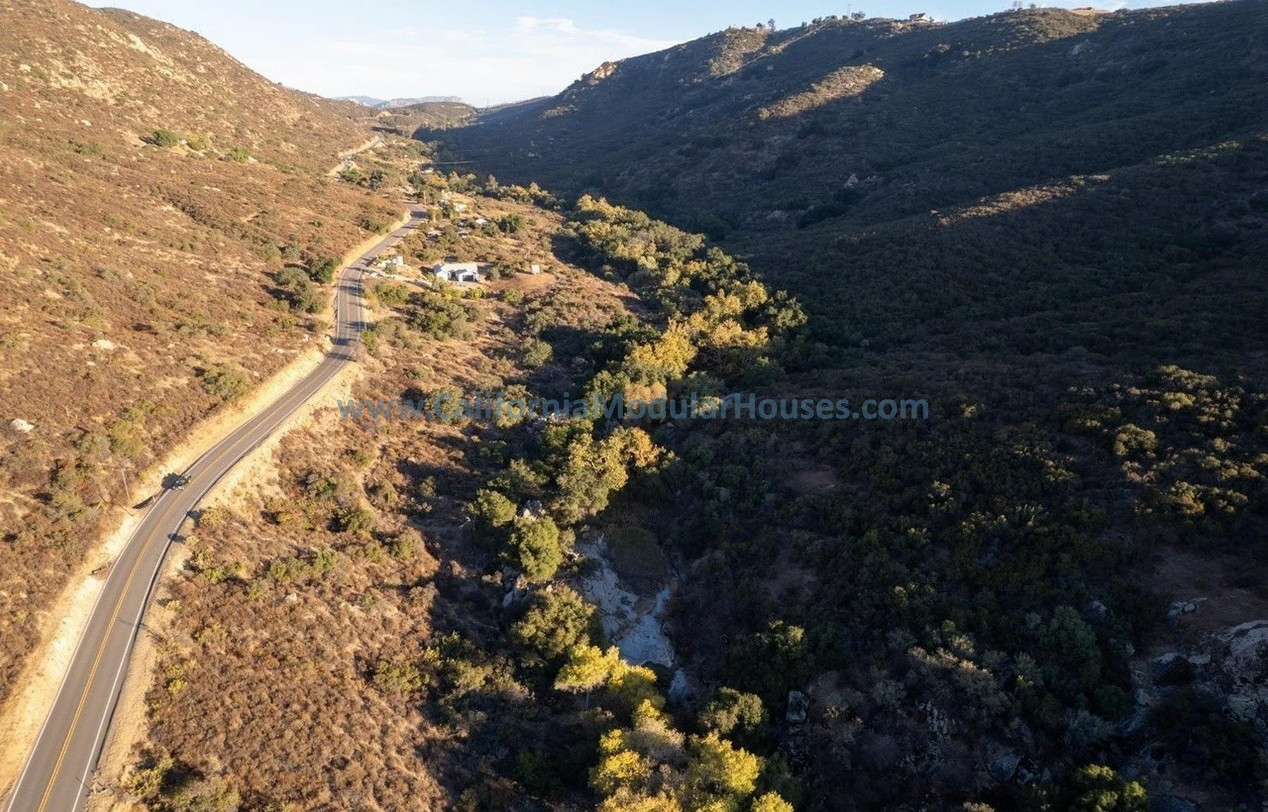 An aerial view of a winding road through a hilly, semi-arid landscape with sparse vegetation and a few small buildings.