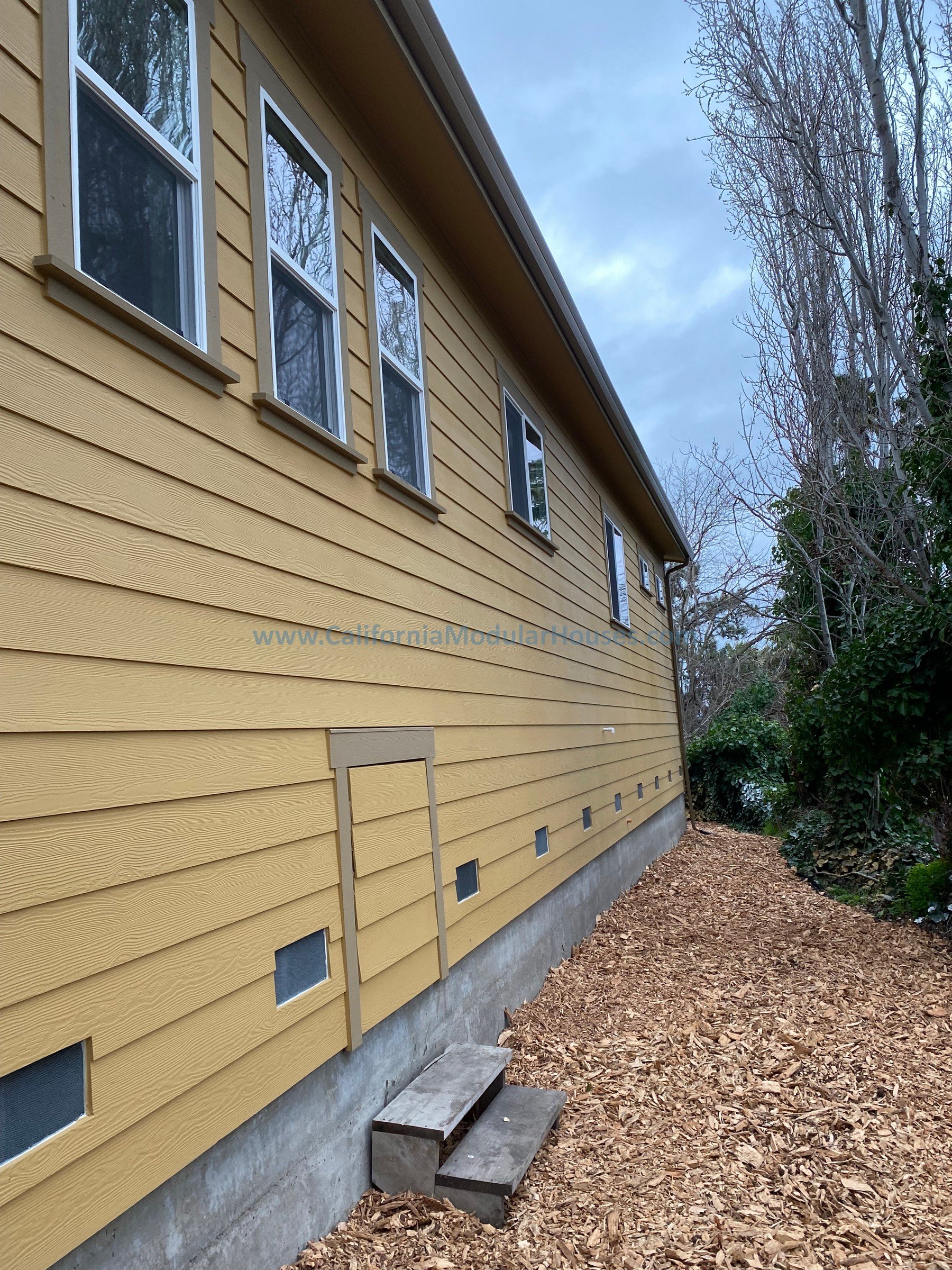 Side view of a yellow house with multiple windows, small outdoor steps, and a wooded yard.  Modular ADU  Prefab modular.  