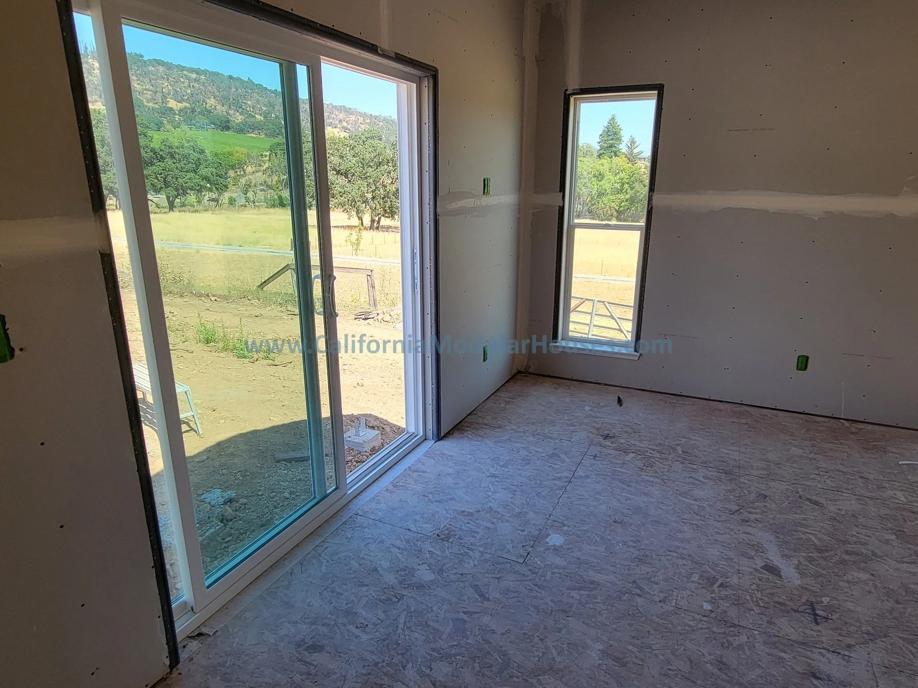 Interior room under construction with sliding glass door and window overlooking a rural landscape with hills, trees, and fields.