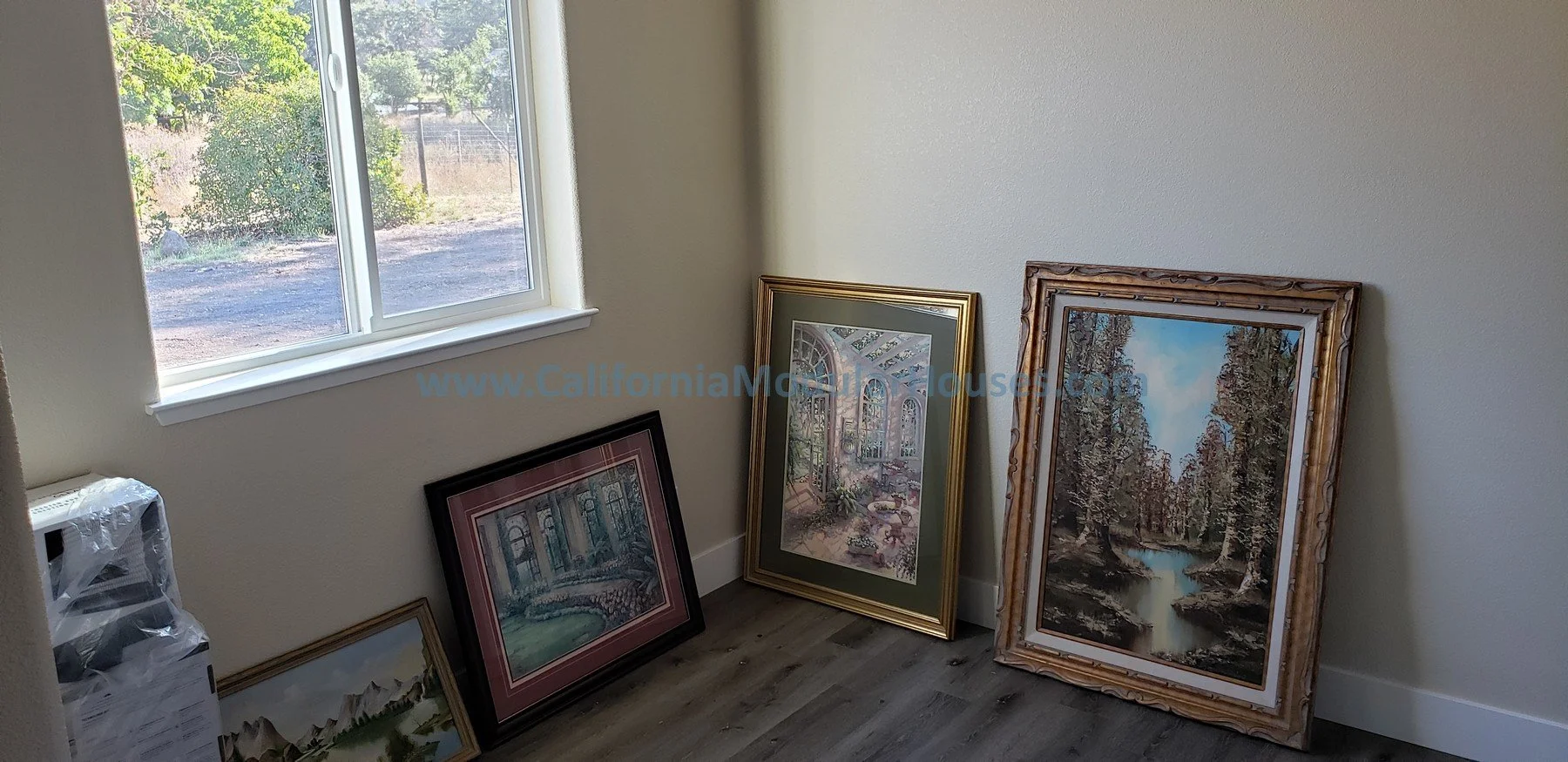A room with light-colored walls and a window, with four framed landscape paintings resting on the floor, showing various nature scenes including mountains, lakes, and trees.   Kenwood, Sonoma County, CA. Backyard home.  