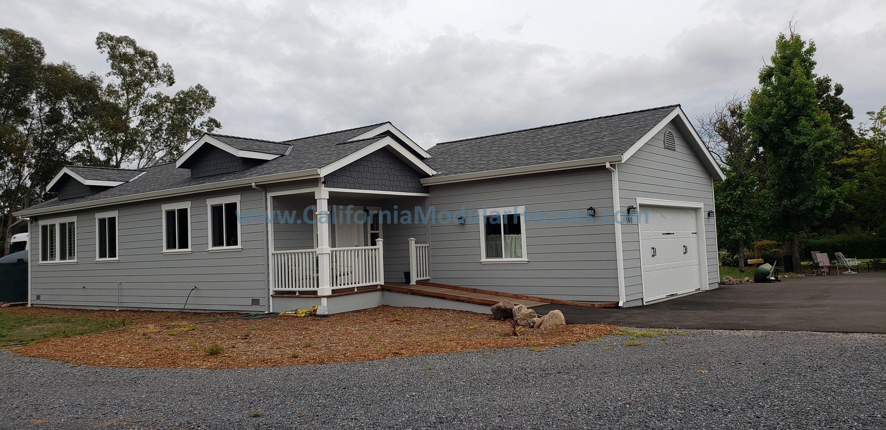 A newly constructed light gray house with a dark gray roof, white trim, and a small covered front porch with white railings. The house has multiple windows and an attached garage with a white door. The driveway is paved, and there are some trees and 