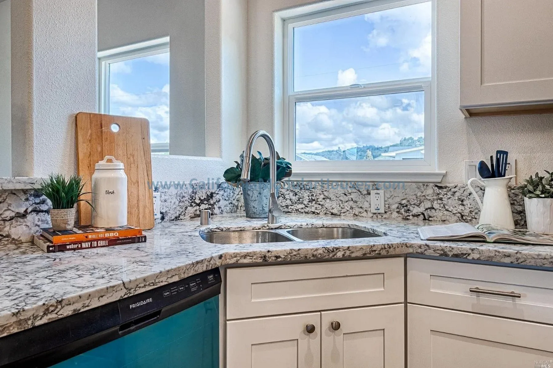 Kitchen sink with double basin, granite countertop, window showing blue sky and clouds, potted plant, books, an open cookbook, and various kitchen utensils.