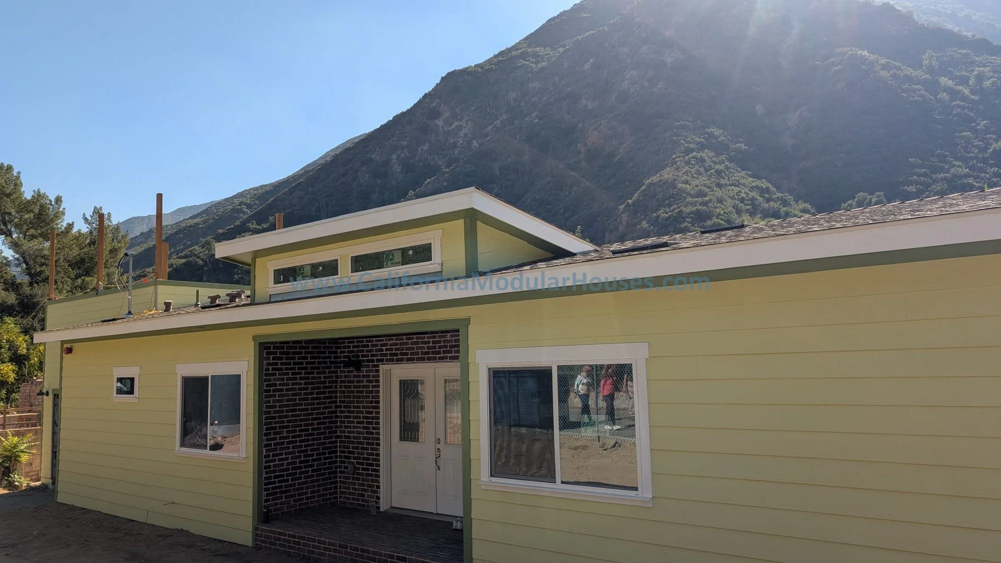 Newly constructed yellow house with white trim and brick entryway, set against a backdrop of mountains and a clear blue sky.