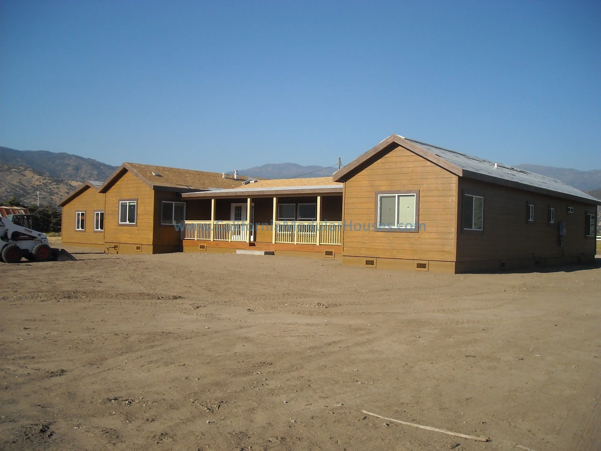 A single-story wooden house with a huge covered porch, with mountains in the background under a clear blue sky.  It's awesome in the winter with snow on the mountains.  Factory Built Housing.  