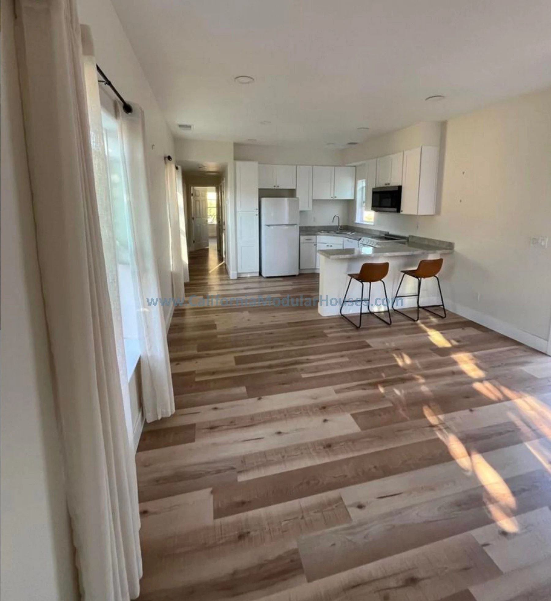 View of a kitchen with white cabinets, a refrigerator, a microwave, and a breakfast bar with two brown chairs, hardwood floors.  Interior view of prefab modular home.  Modular Home CA.  Oceanside, San Diego County, CA.    