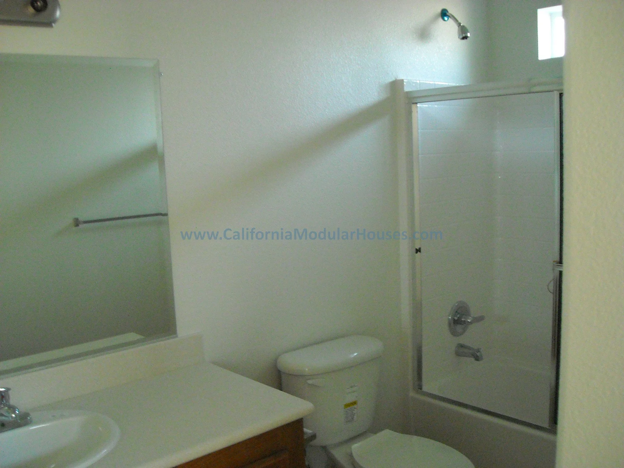 Bathroom with a sink, mirror, toilet, and a shower with a glass door, small window, and beige walls.  Prefab home for the Golden State of California.  
