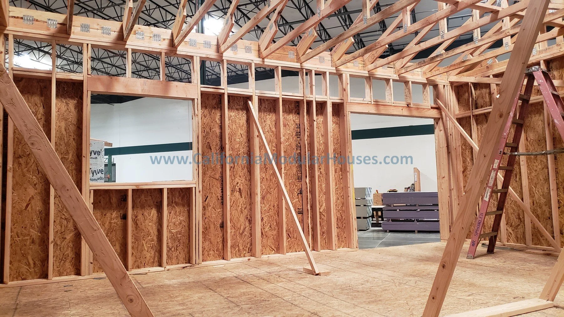 Interior view of a building under construction with exposed wooden framing and OSB wall sheathing, a red ladder leaning against the framing, and a large open doorway leading to another room.