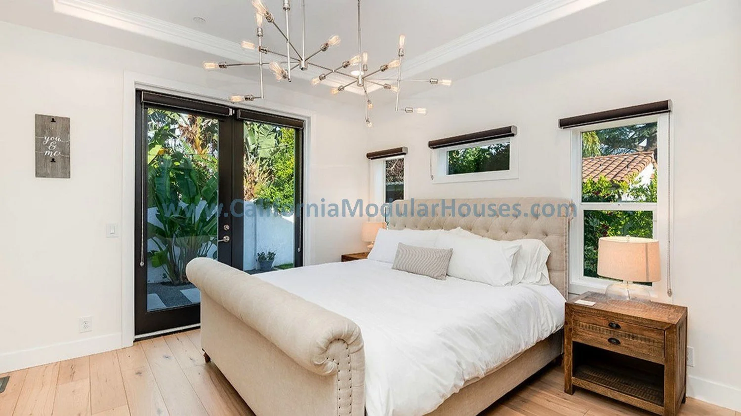 A modern bedroom with white walls.  There are three windows behind the bed and a glass door leading to a patio with greenery outside. A contemporary chandelier hangs from the ceiling. This shows a coffered - trey ceiling.  
