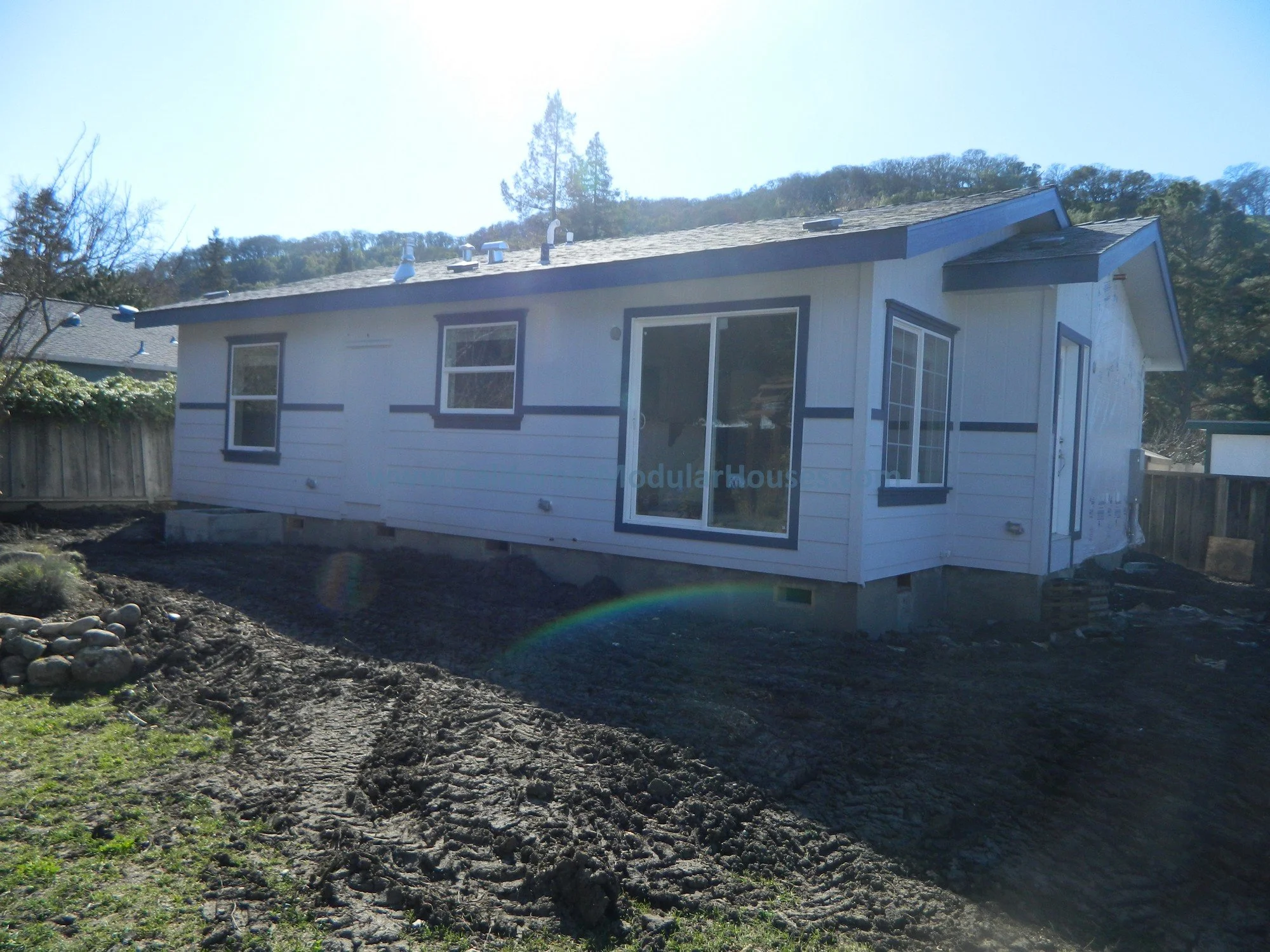 A small, white prefabricated house under construction, elevated on concrete blocks, with several windows, set on a dirt lot with tire tracks and patches of grass, surrounded by a wooden fence, with a hillside covered in trees in the background.
