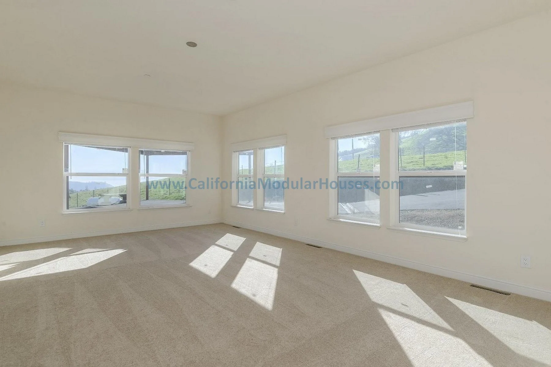 Empty room with beige carpet and large windows letting in sunlight, showing a green hilly landscape outside.  Bay Area Prefab Homes, Modular Homes California, Modular Homes,