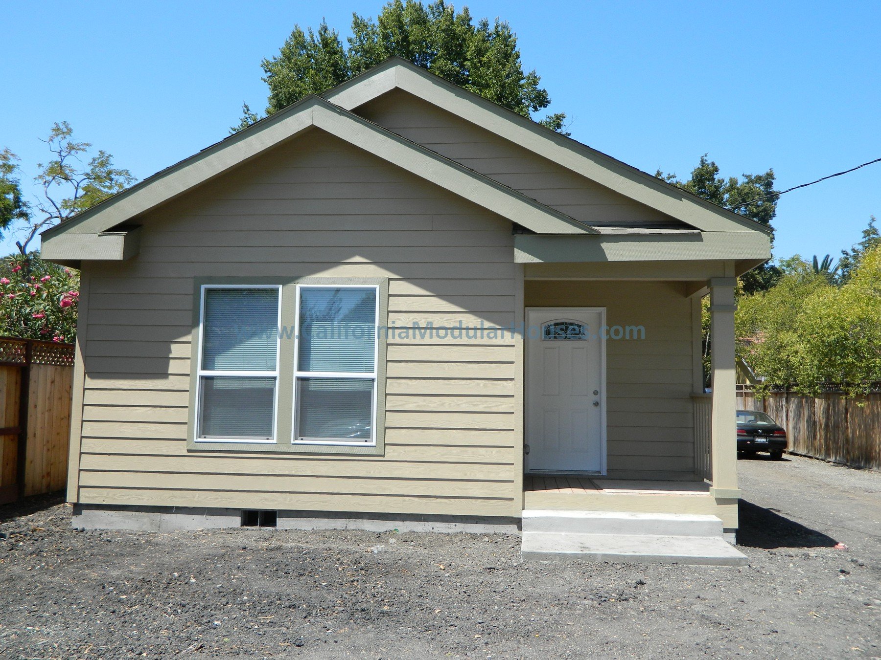 Front view of a small beige house with two windows, a white door, and a small covered porch with concrete steps, in a yard with a gravel driveway, wooden fence, and trees in the background.