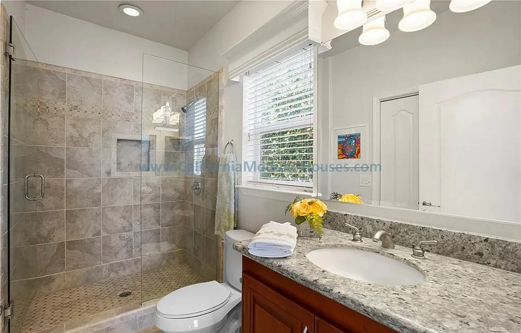 Bathroom with a walk-in shower enclosed with glass, a toilet, a granite countertop with a sink, window with white blinds, and a white door. California Modular Houses. Modular home construction California.  Prefab house.  Oxnard, Ventura County, CA.  