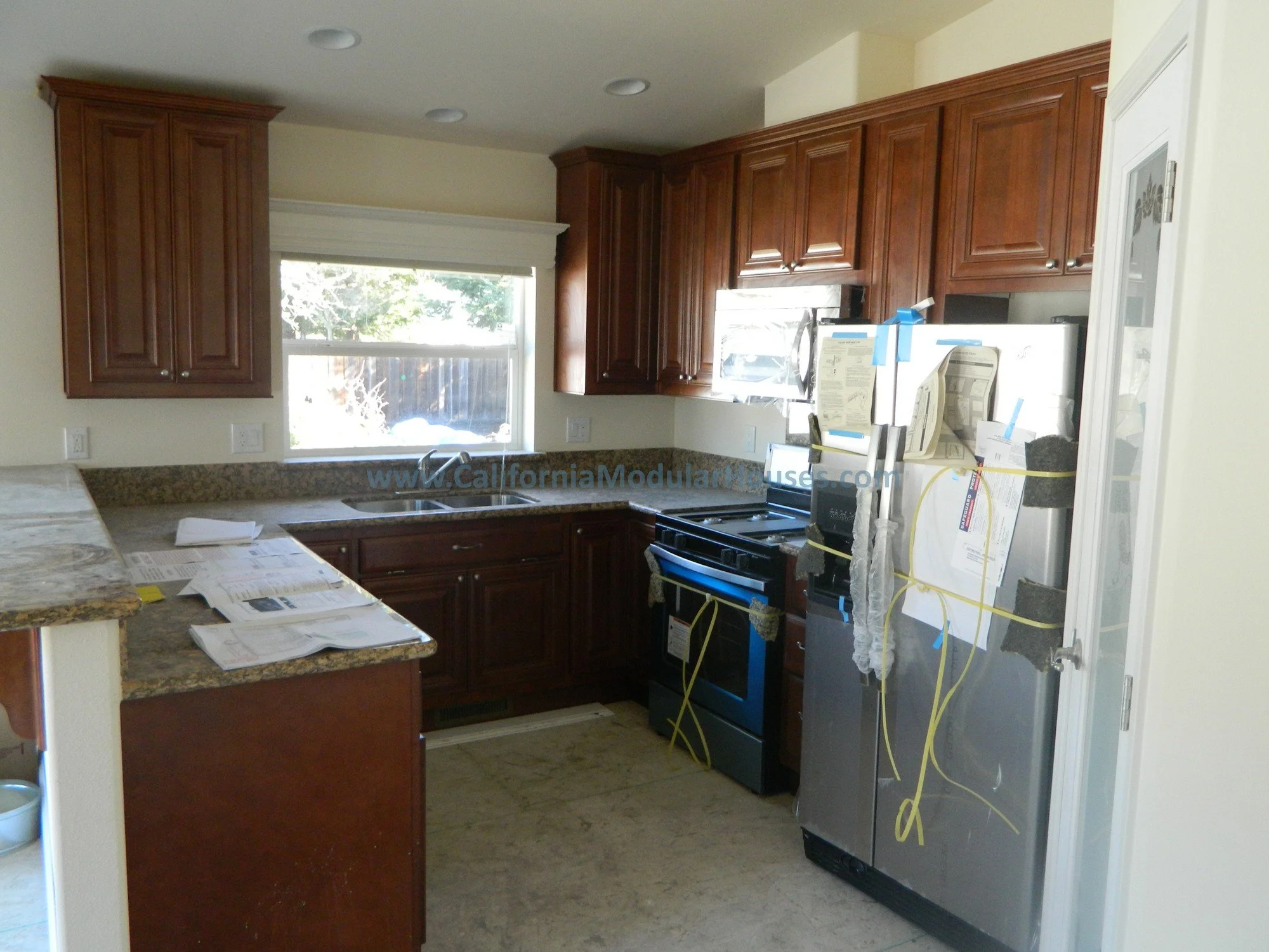 Kitchen with wooden cabinets, granite countertops, a window above the sink, and a stainless steel refrigerator covered in papers and zip ties.