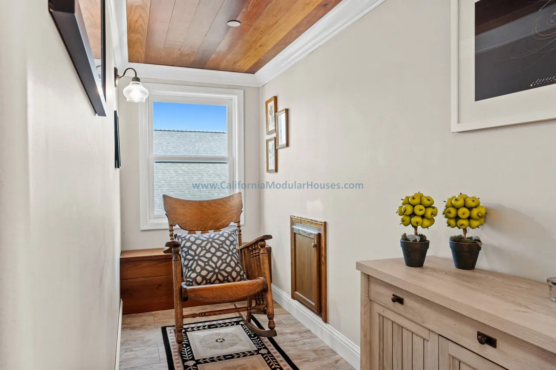 A small cozy room with a wooden chair, a window letting in natural light, a wooden side table with yellow apple-shaped decorations, framed artwork on the beige wall, and a wooden ceiling with a wall-mounted light fixture.  Prefab modular home.  CA.  