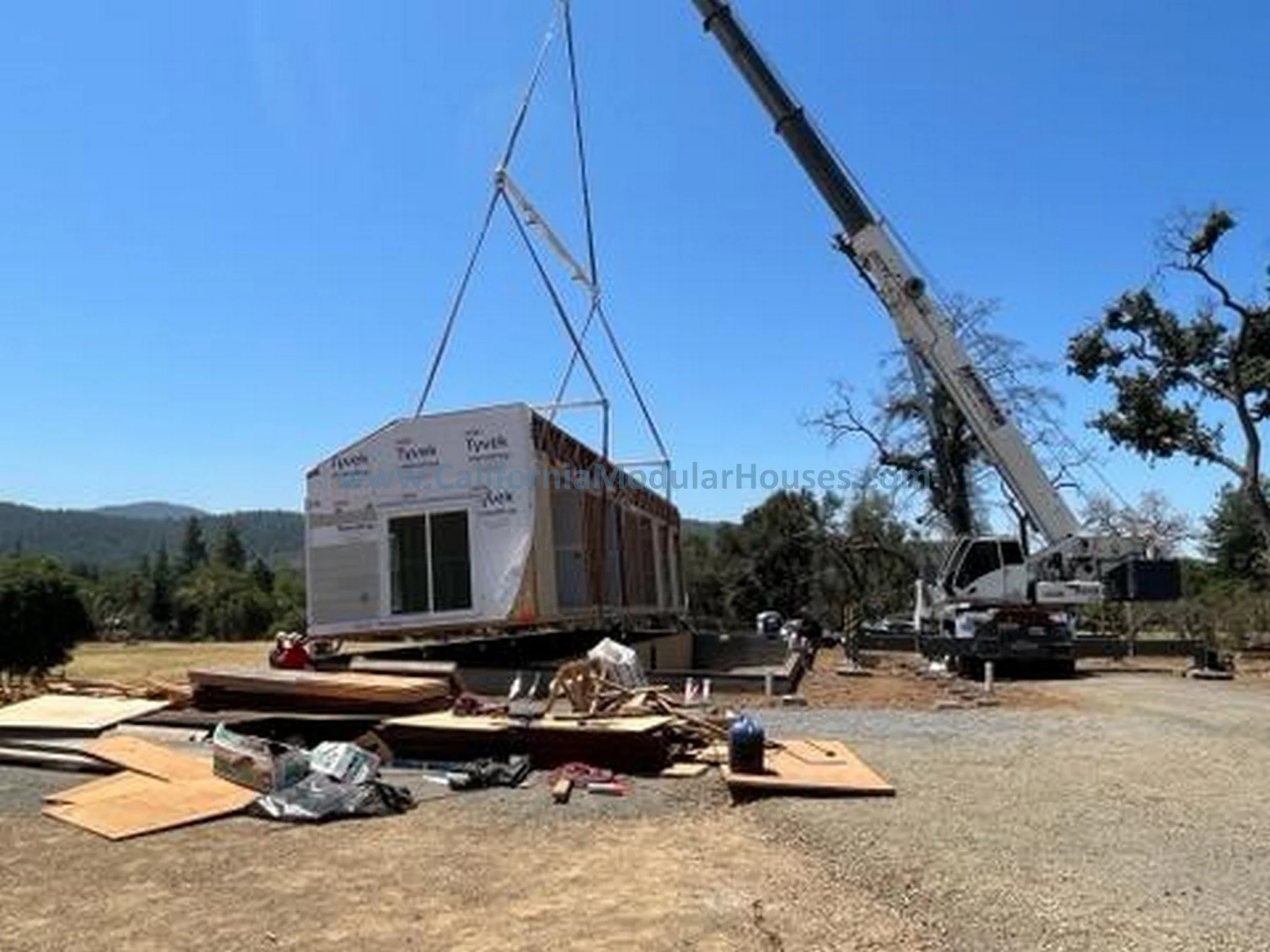 A house under construction being lifted by a crane on a construction site with various construction materials on the ground.
