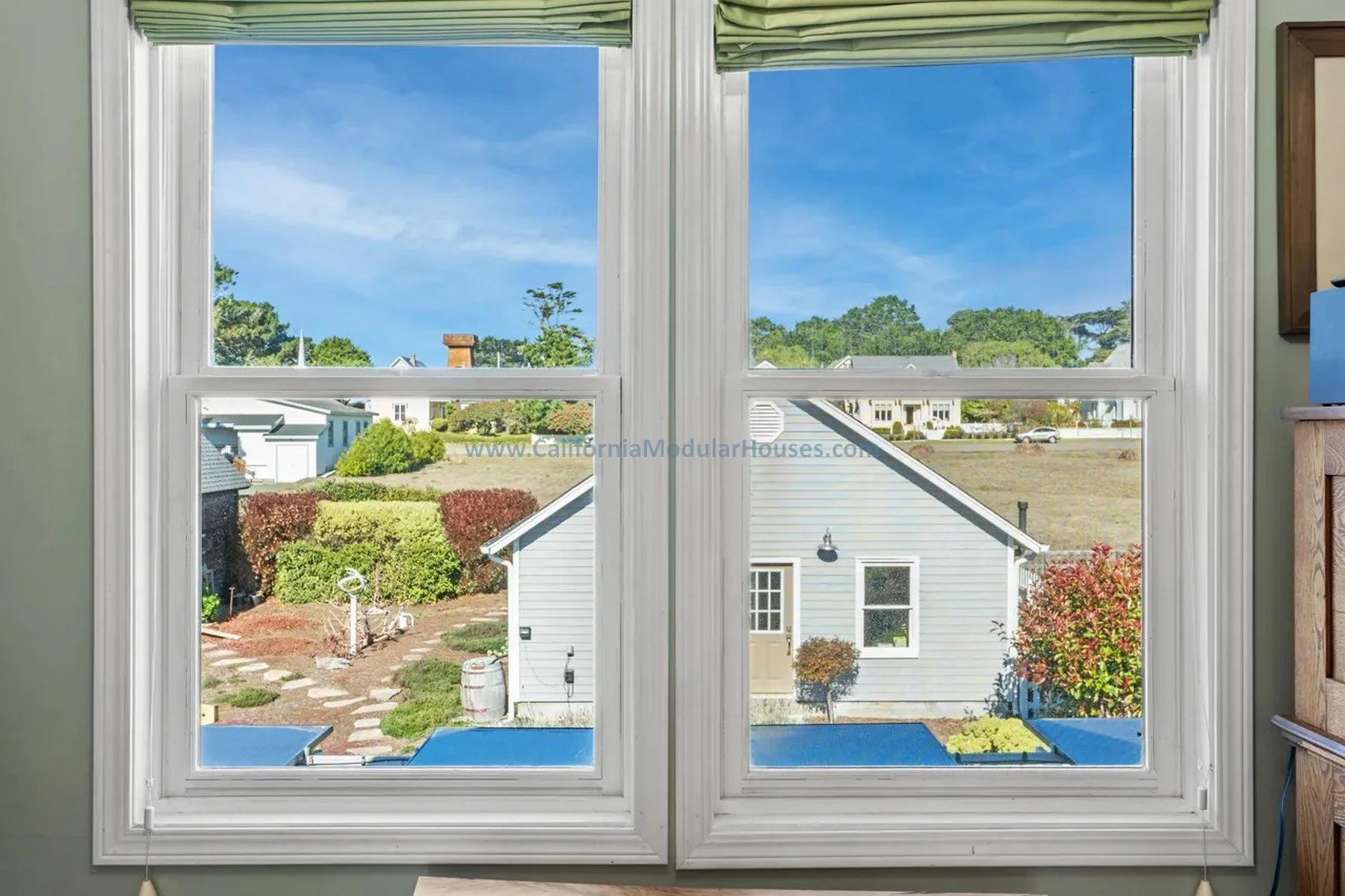 View through a large window showing neighboring houses, greenery, blue sky, trees, bushes, and a small garden with stepping stones. Historical design. Mendocino City, CA. Factory Built Home.  Mendocino County, California.