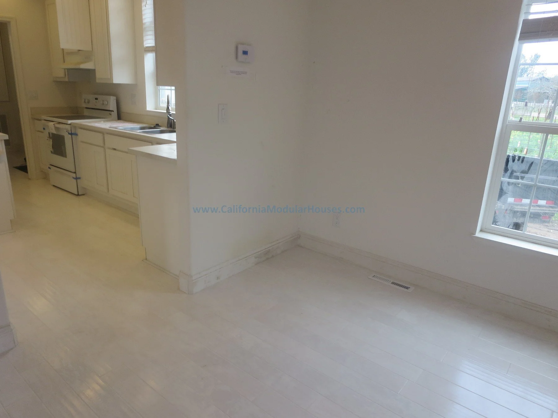 Empty room with white walls and light-colored hardwood floors, adjoining a kitchen. The room has large windows allowing natural light. Napa County, CA Vineyard.  Modular homes.  SFR.  New House Cape Cod.  Unfinished attic.  