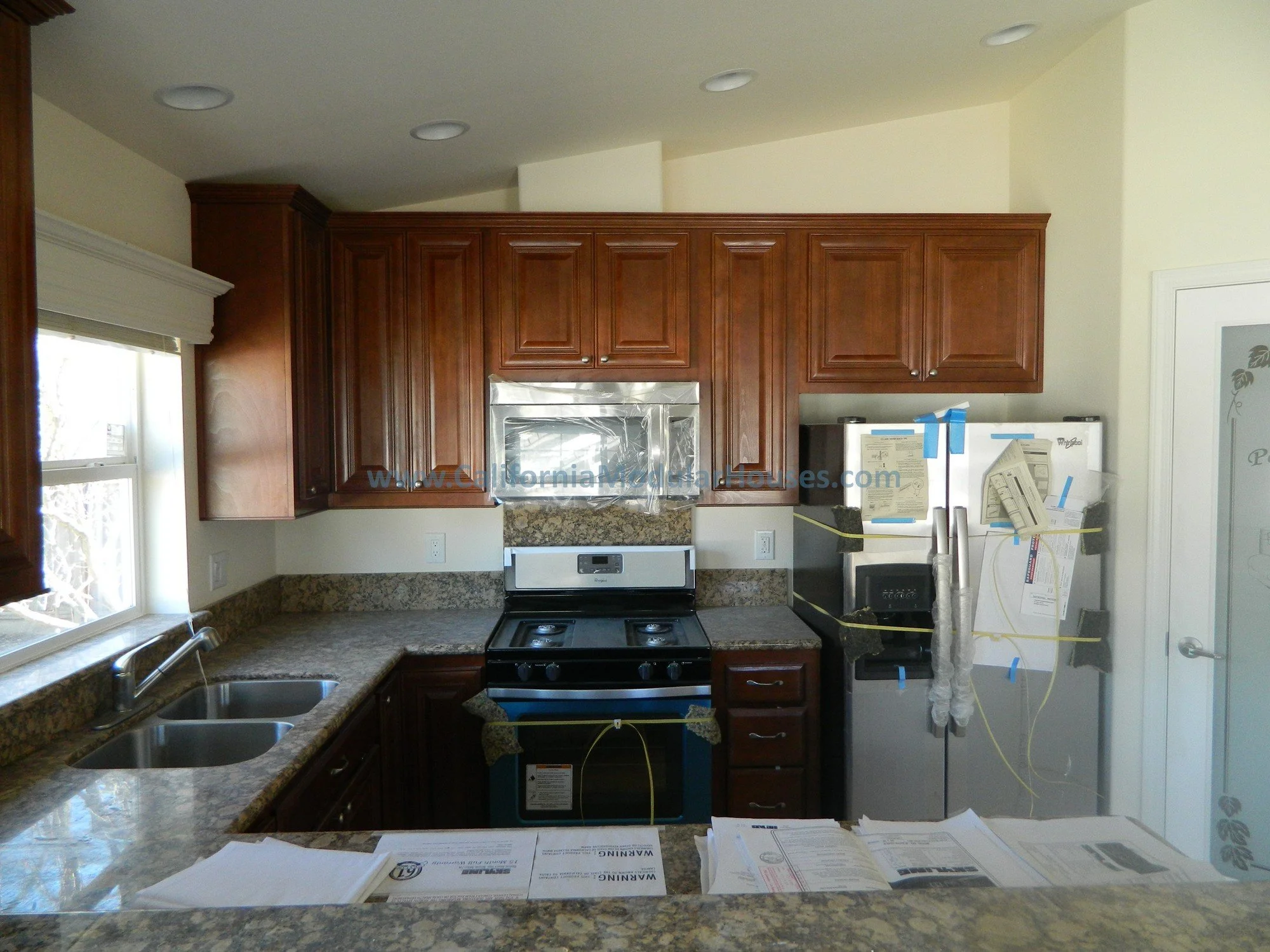 Kitchen with wooden cabinets, granite countertops, stainless steel microwave over stove, and a refrigerator with blue painter's tape and papers attached.