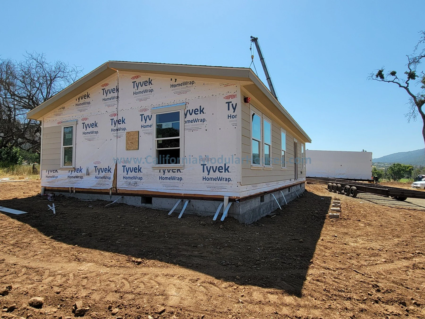 A house under construction with exterior walls partially covered in Tyvek HomeWrap. The house is elevated on a foundation with exposed supports, and there is a crane nearby lifting construction materials. The surrounding area is a dirt lot with some 