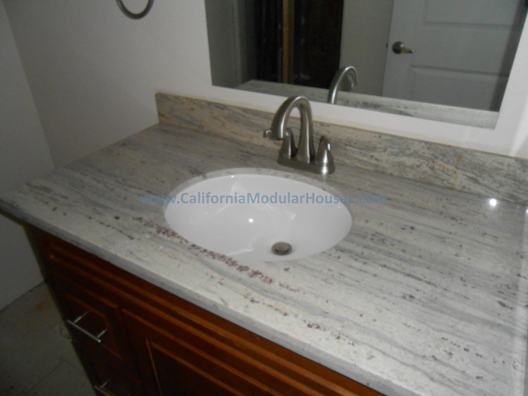 Bathroom vanity with a granite countertop, an inset oval sink, and a mirrored wall above. The faucet is brushed nickel, and part of a white door and a wall are visible in the background.  Modular Home CA, California Modular, Modular Homes,