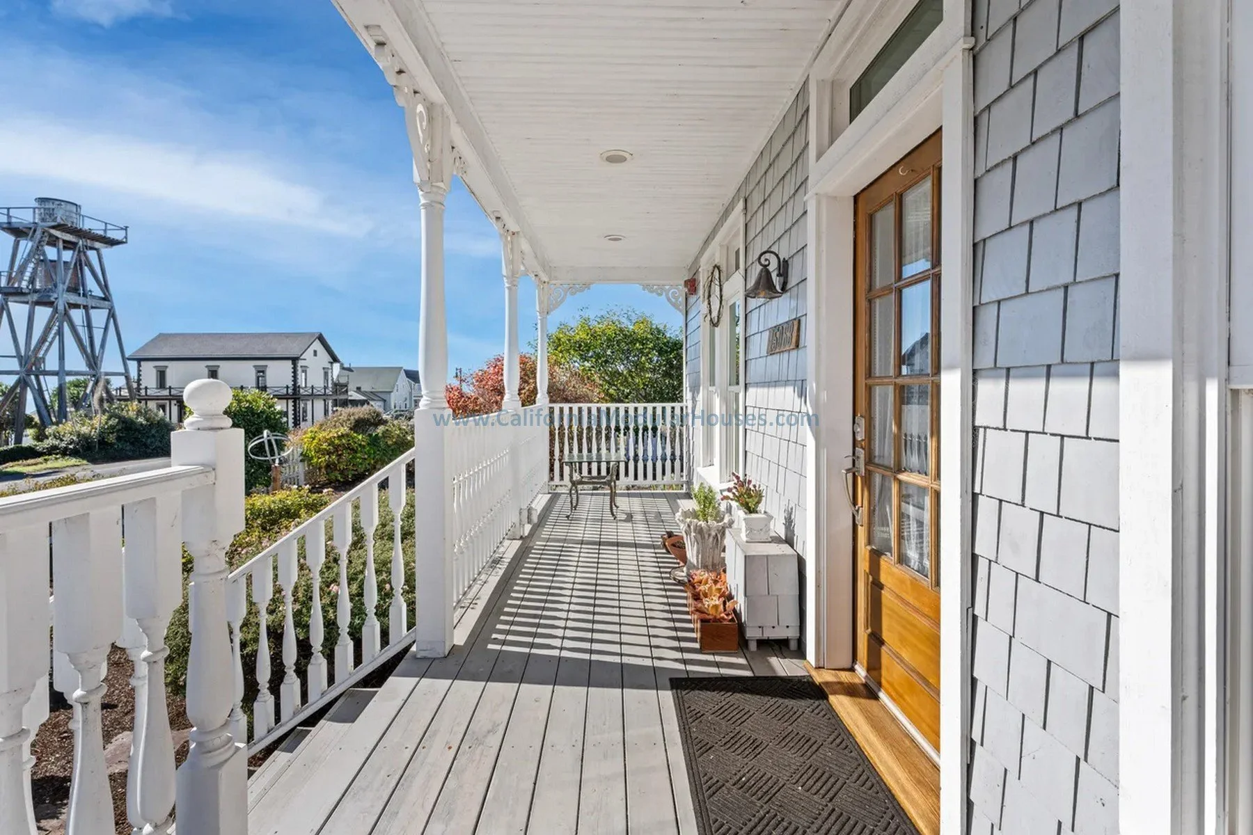 Front porch of a house with white railing, wooden floor, and a wooden door with glass panes. The porch has potted plants and a small table with chairs. Mendocino City, CA.  Mendocino County, California.  Prefab modular.  Historical.  