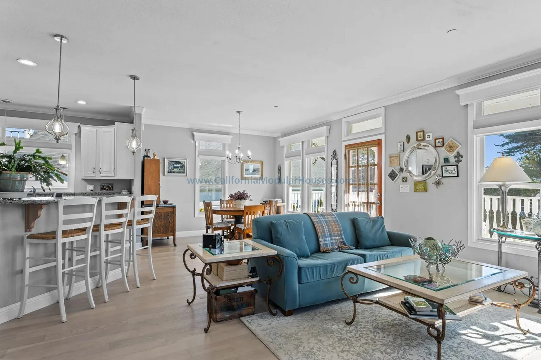 Living room with a blue sofa, glass-topped coffee table, decorative wall mirror, and multiple windows letting in natural light. Visible dining area in the background.  Historical. Mendocino City, CA. Prefab modular. Mendocino County, California. 
