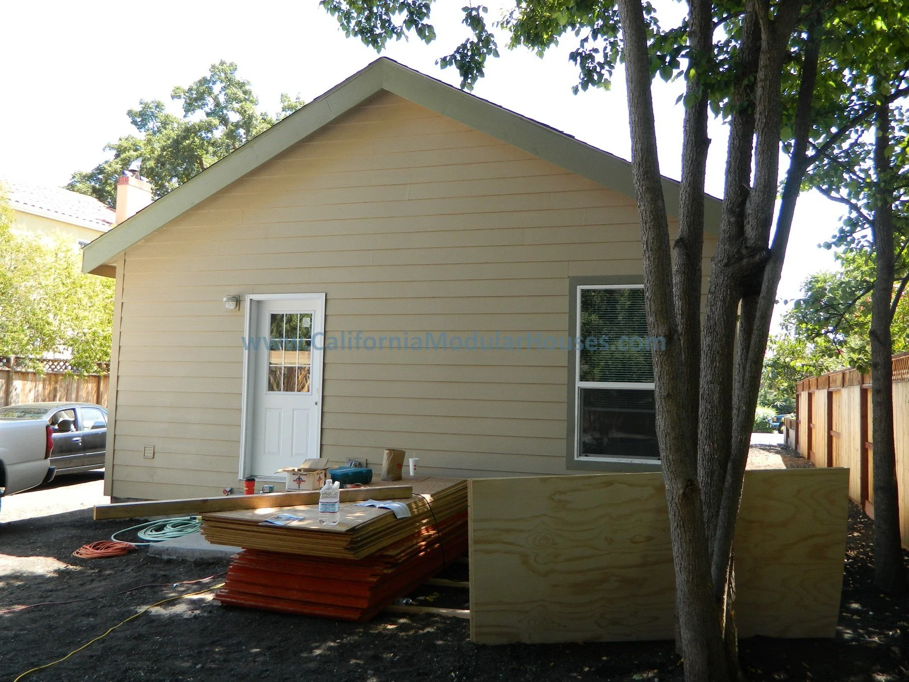 Construction of a small beige house with a gabled roof, with building materials and tools on the ground in front, and a white door and window visible.