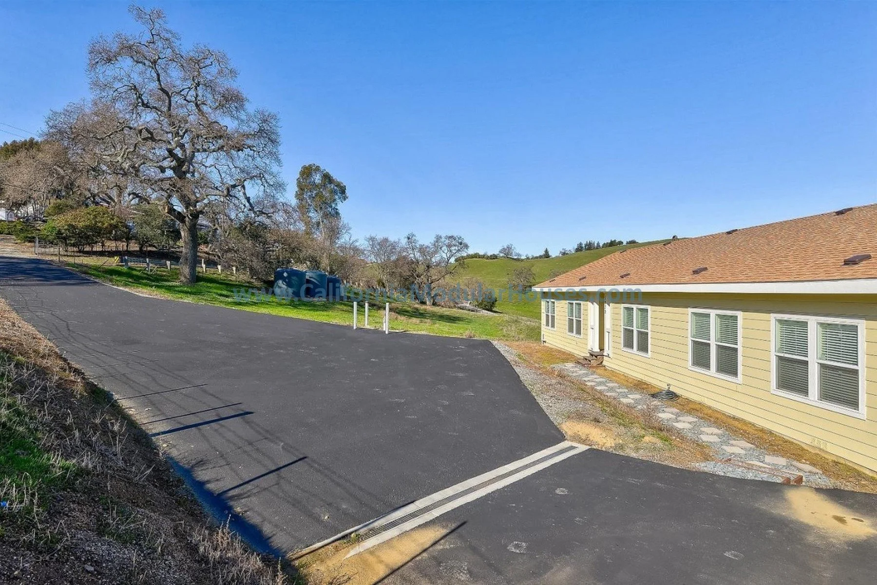 A residential area with a yellow house, a paved driveway, a large leafless tree, green hills in the background, and clear blue sky.  California Modular Houses, Bay Area Prefab Homes, Pre-Fabricated Homes,