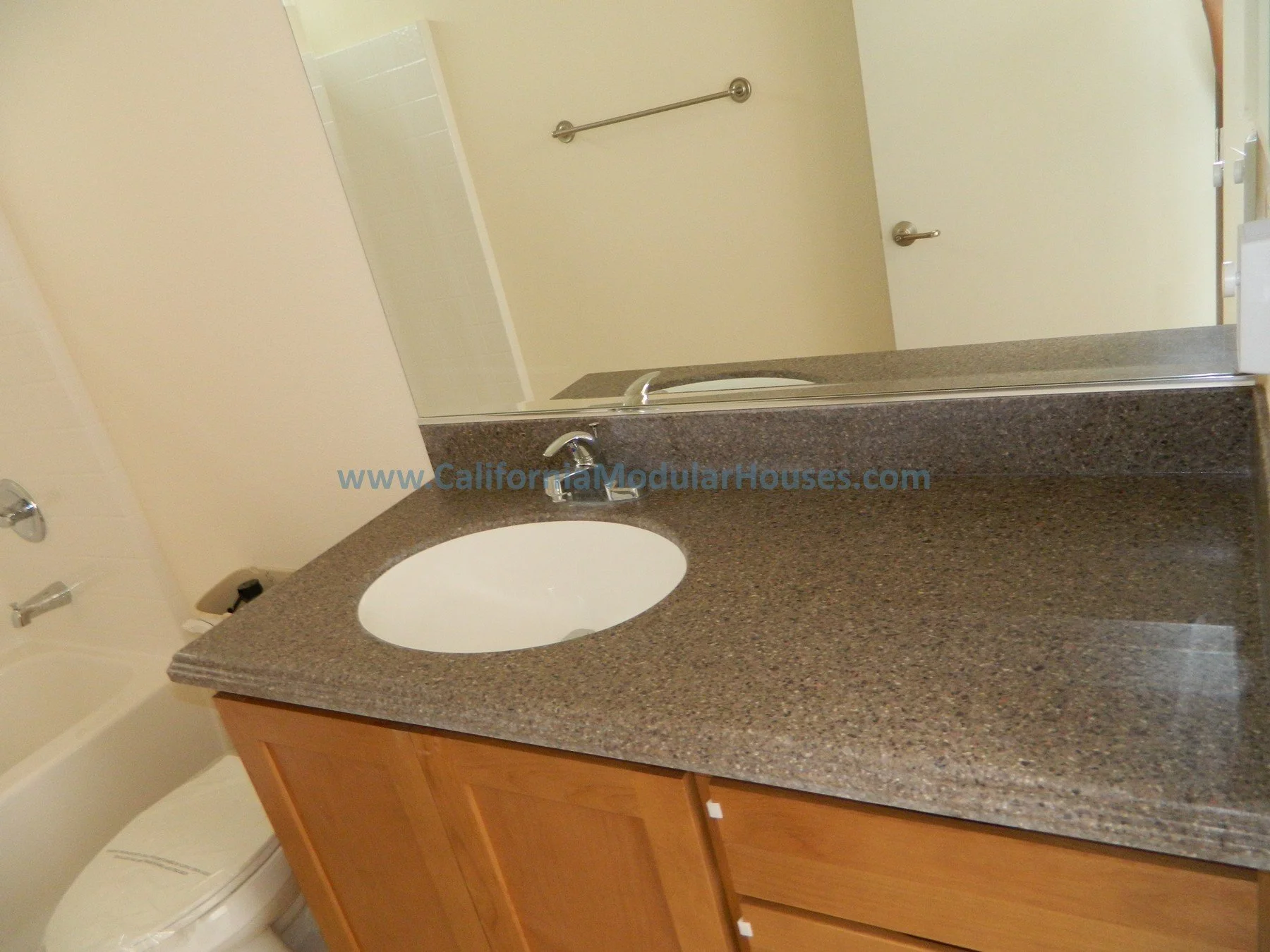 Bathroom with a granite countertop, oval sink, mirror, wooden cabinet below the sink, and bathroom fixtures including a soap holder, towel bar, and toilet. Northern CA Modular Home, California Prefab Modular Home.