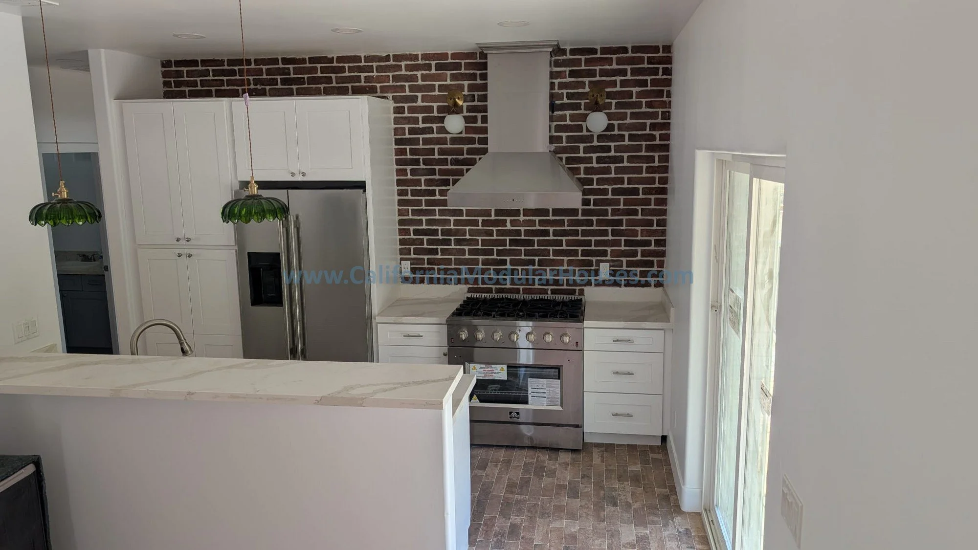Kitchen area with white cabinets, a stainless steel refrigerator, a gas stove, a stainless steel range hood, brick accent wall, two green pendant lights, and a sliding glass door.