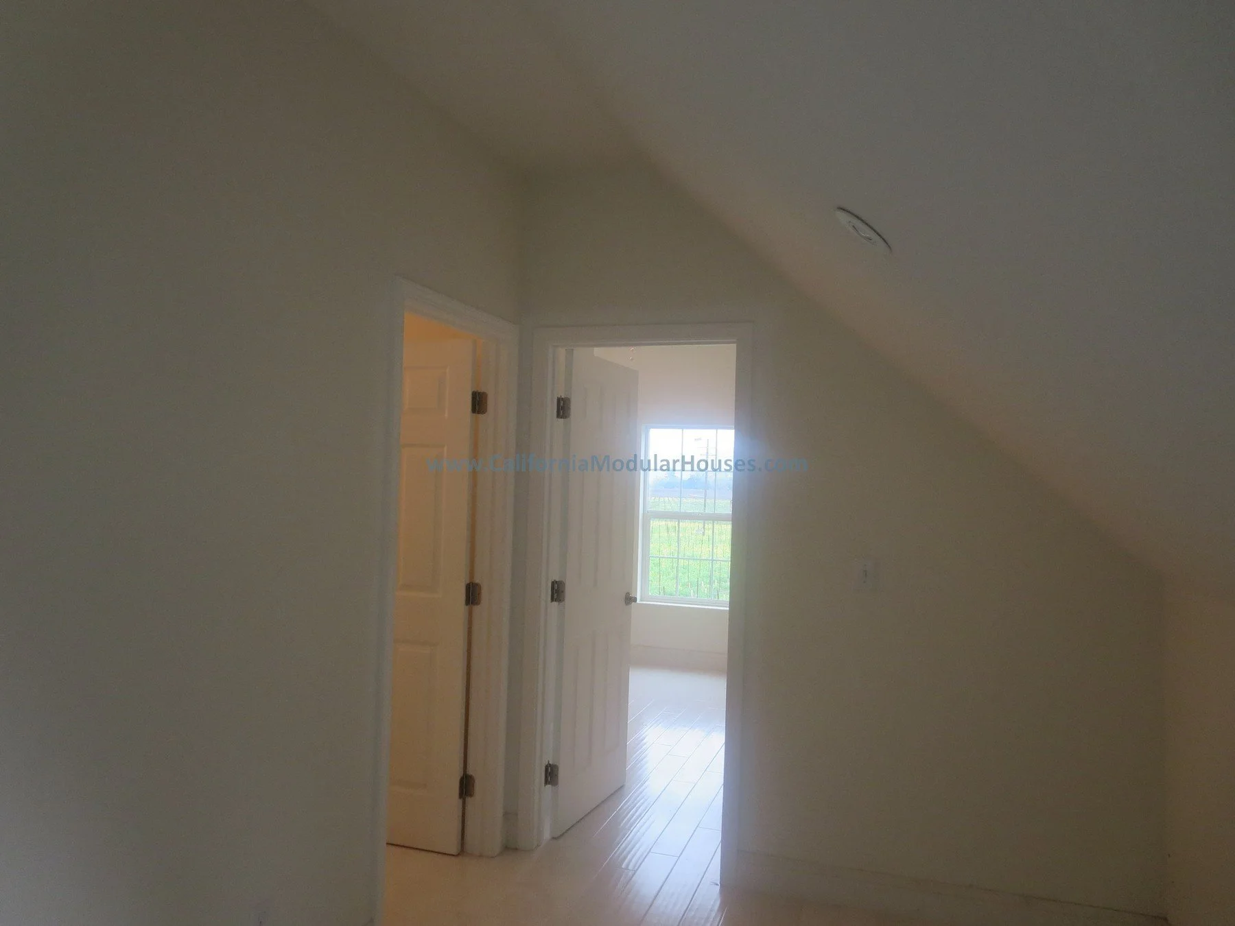 California modular home.  New pre-fab modular house in Napa, California.   Unfinished attic.  Empty room with a sloped ceiling, two open doors leading to another room with a large window, and light-colored walls and flooring.